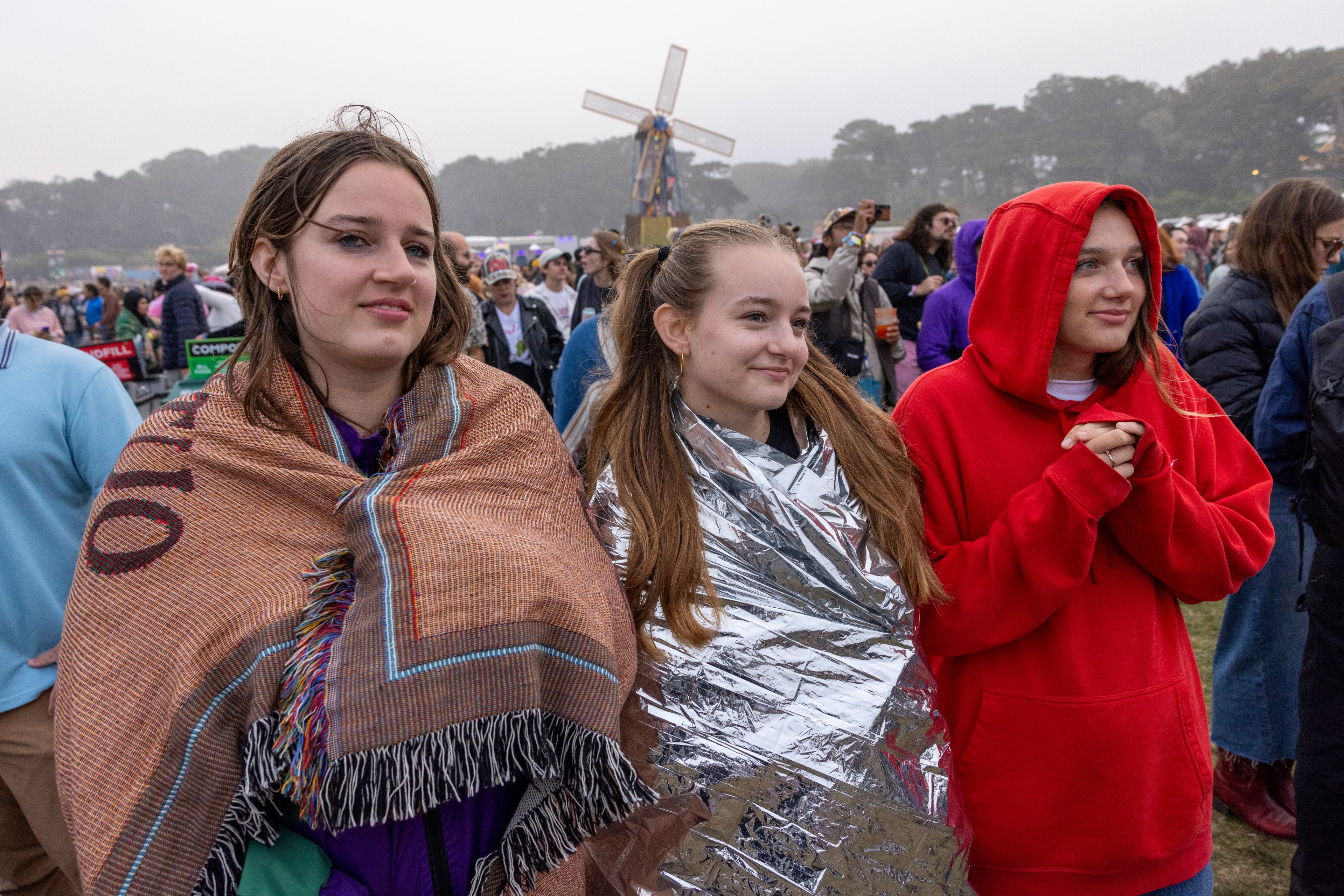 Three young women are wrapped in blankets and a jacket, standing in a crowd outdoors. One wears a brown blanket, another a foil one, and the third a red hoodie.