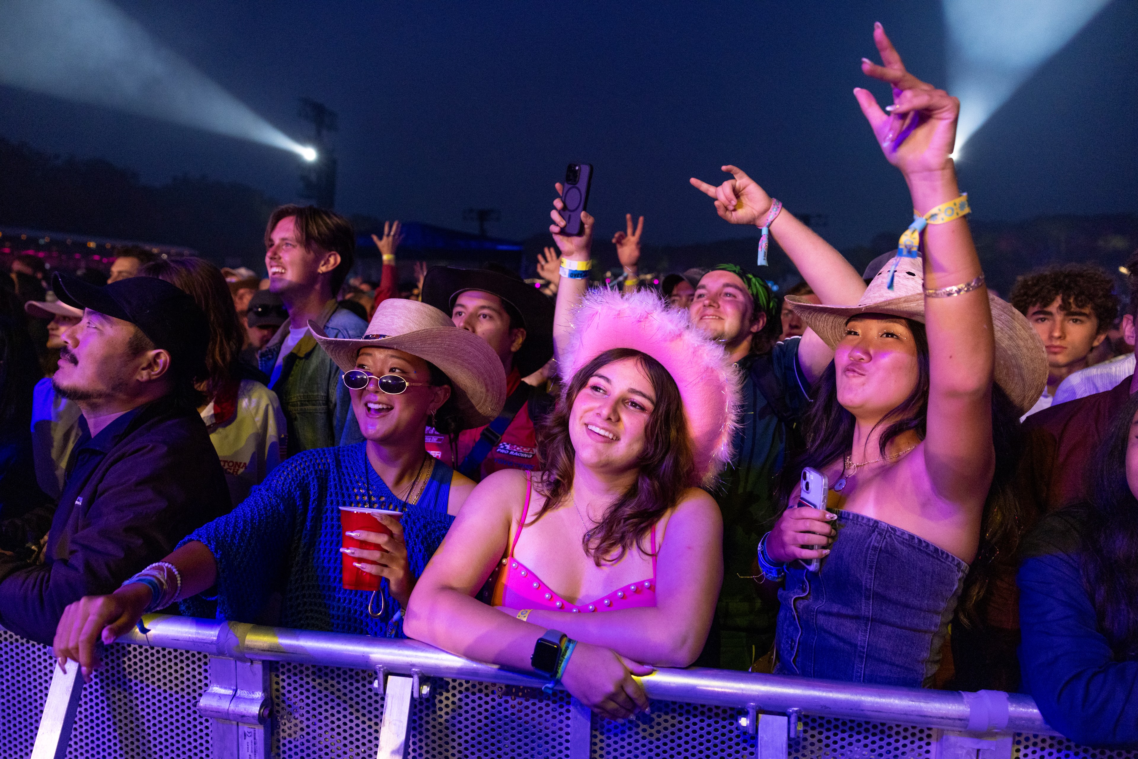 A lively crowd at an outdoor concert enjoys the music. People in colorful outfits and hats are cheering, smiling, and raising their hands under bright stage lights.