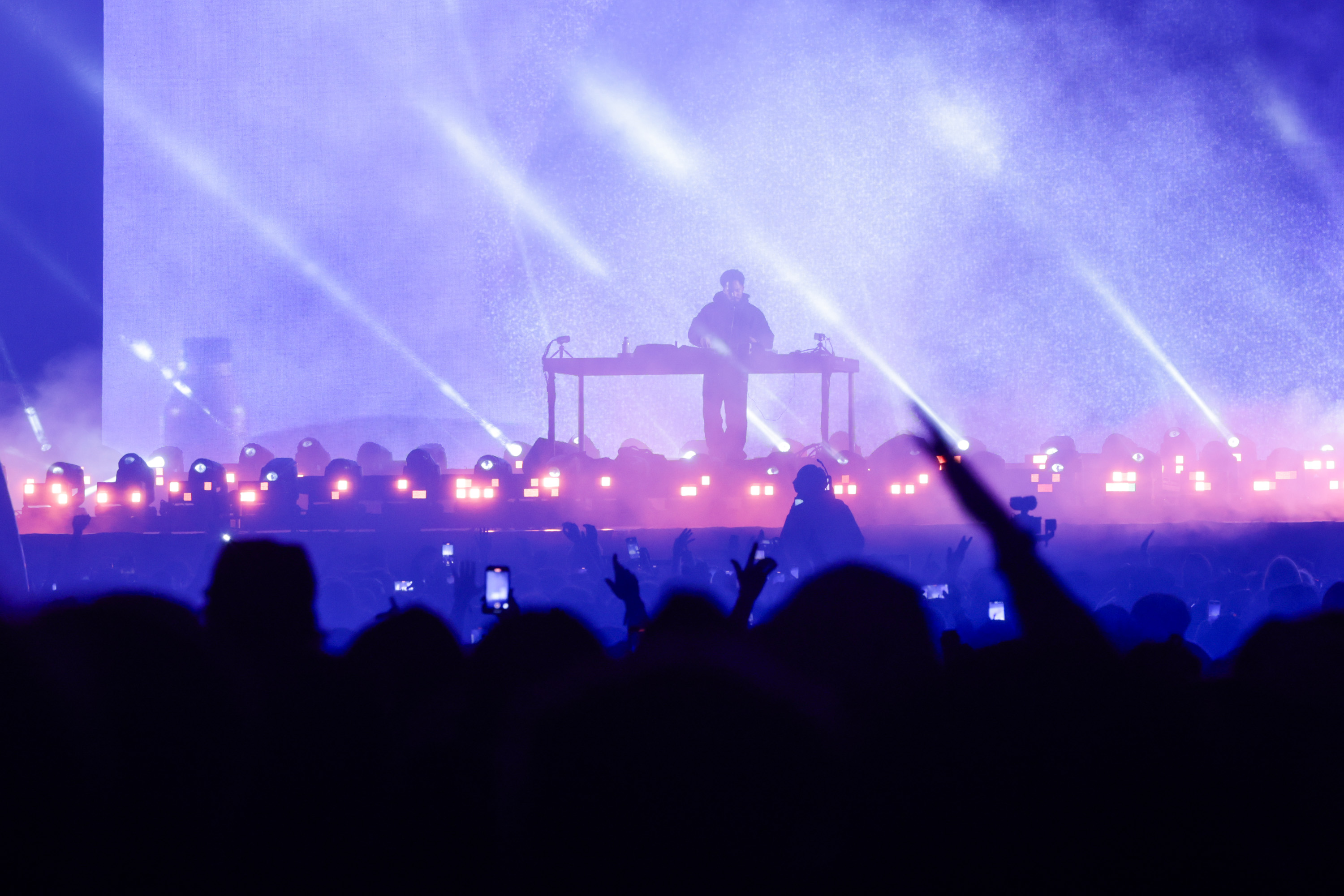 A DJ stands on a stage with vibrant purple and blue lights, surrounded by fog. The crowd below is silhouetted, some holding up phones and cheering.