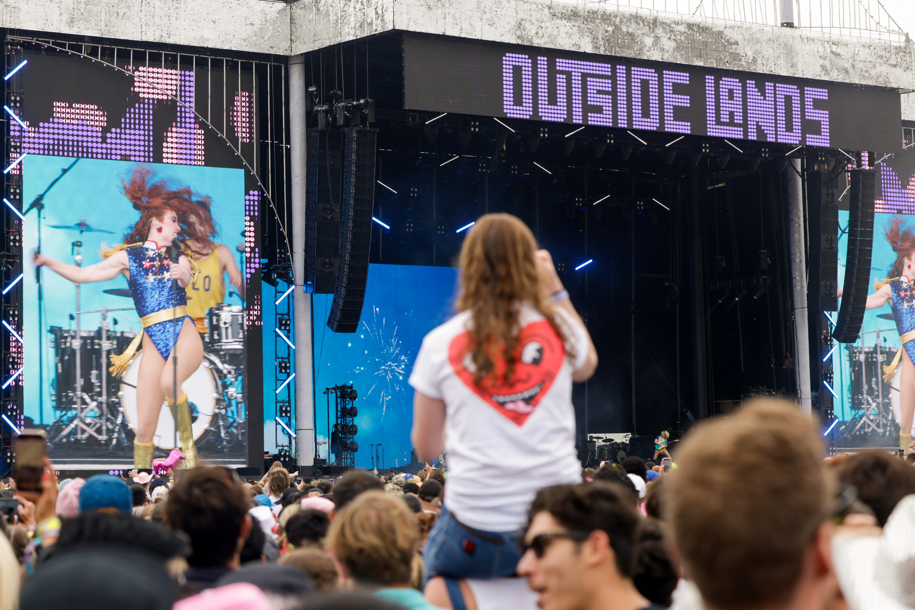 A vibrant concert scene shows a performer in a blue outfit, large screens with her image, and an audience with a person on someone's shoulders under &quot;OUTSIDE LANDS&quot; signage.