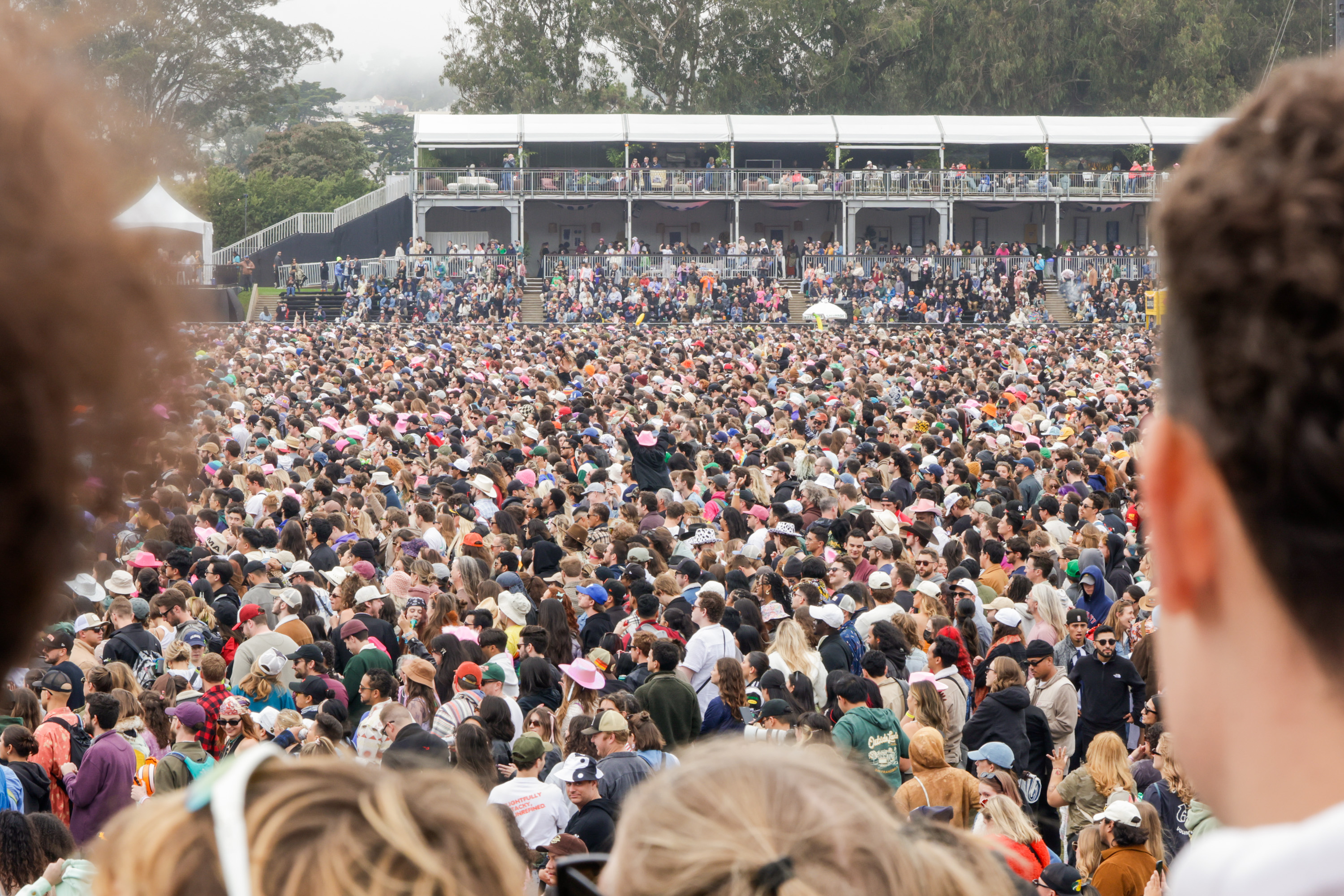 A massive crowd gathers outdoors, many wearing hats, with a large viewing platform and trees in the background. The scene suggests a busy event or concert.