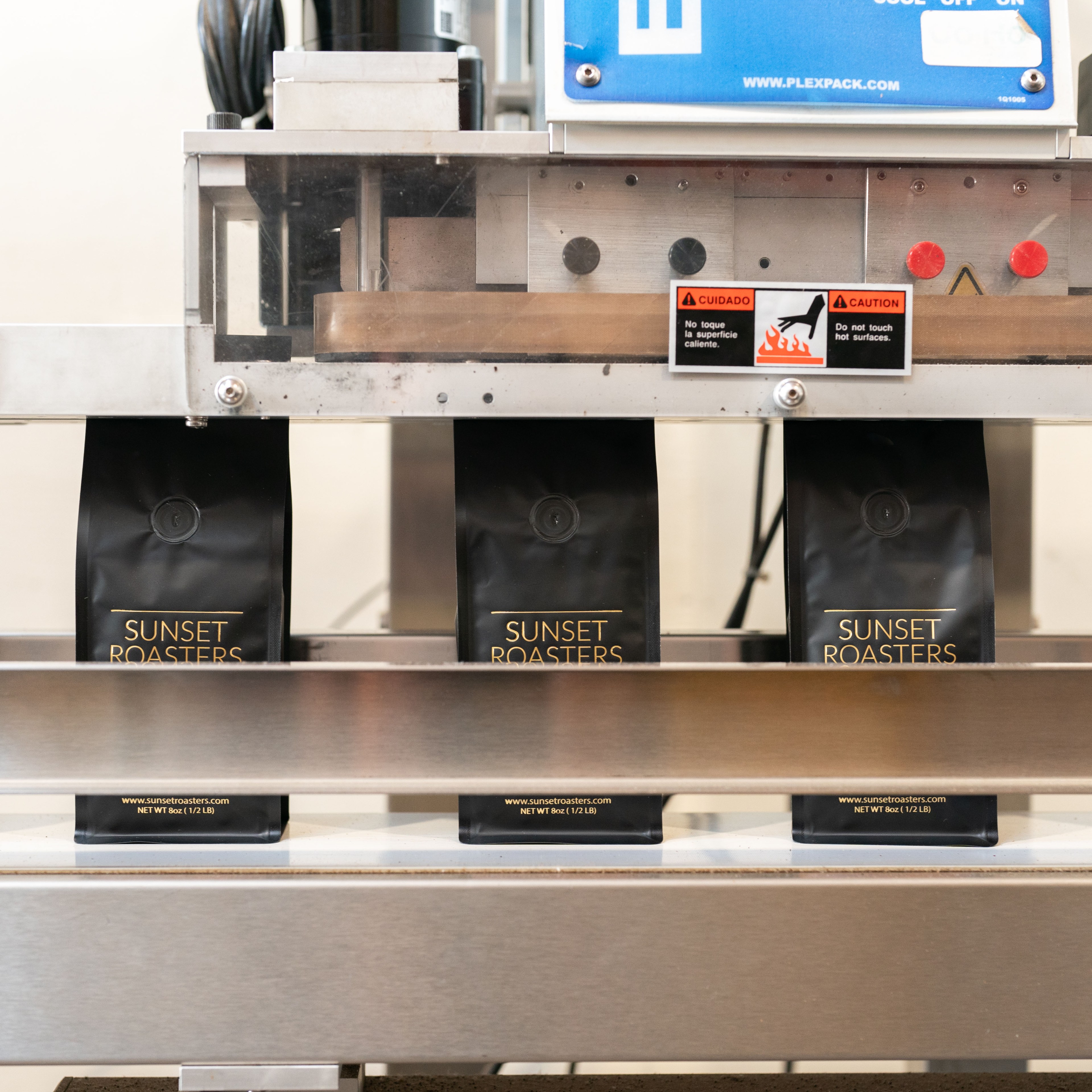 Three black bags with &quot;Sunset Roasters&quot; and a website printed in gold text are being processed under a machine in a factory setting.