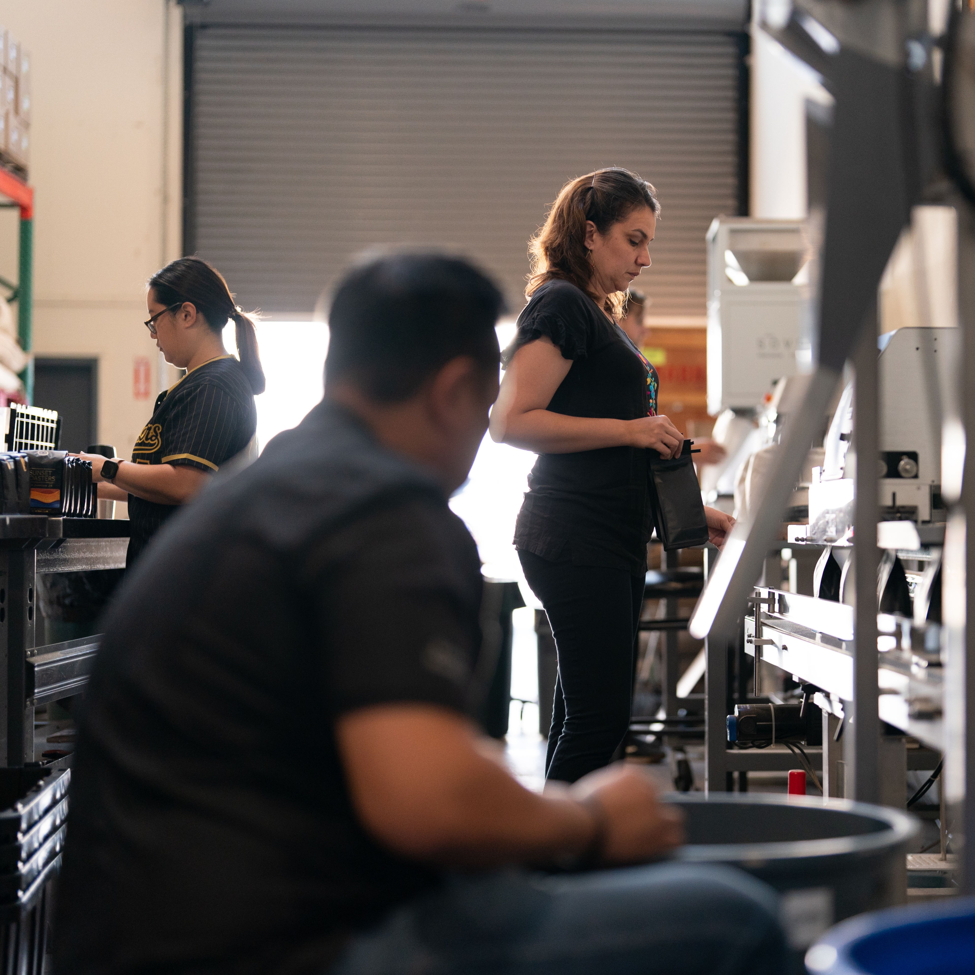 Three people are working in an industrial or workshop setting, focused on their tasks. There is machinery around them and the area is lit naturally from a large open garage door.