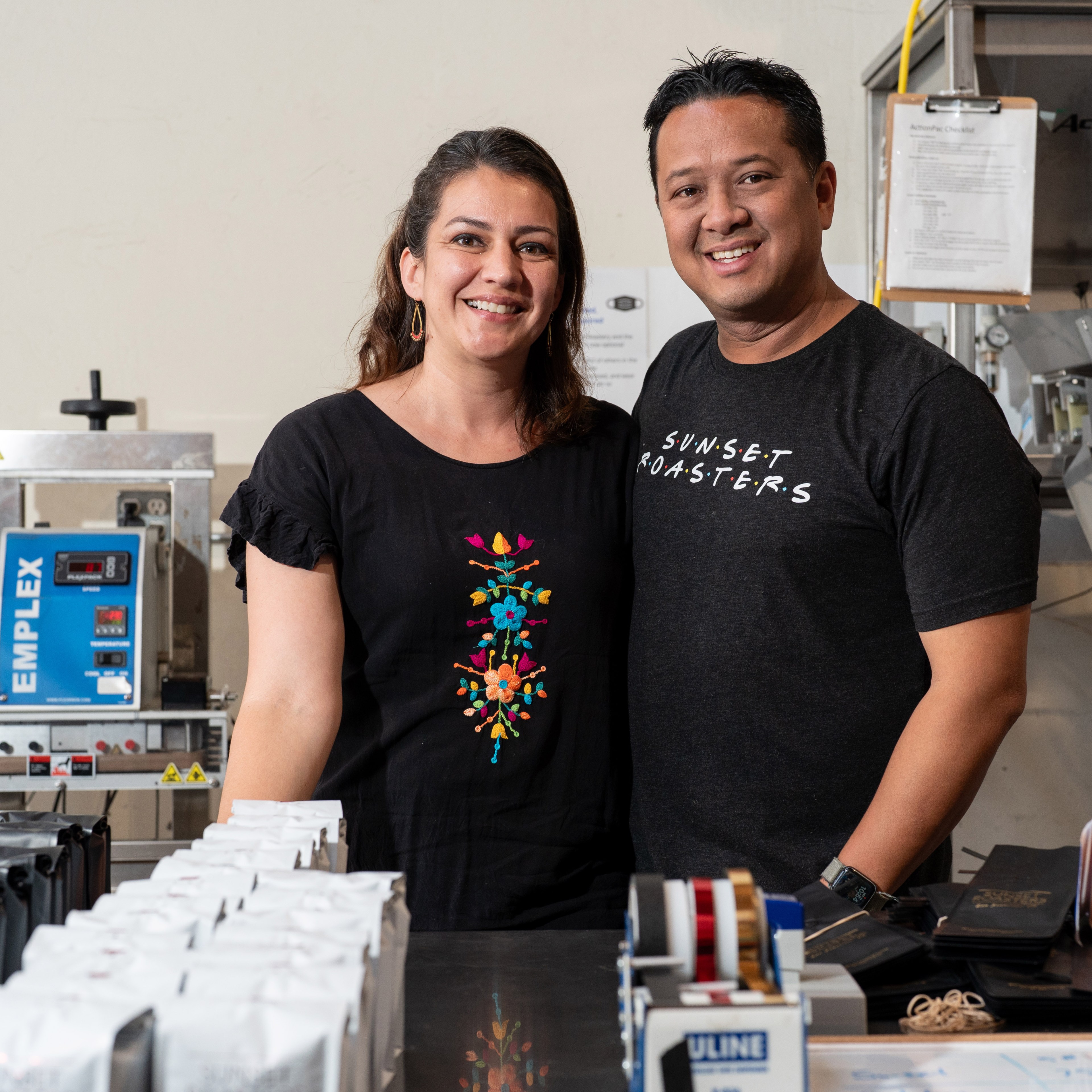 A smiling couple stands in a workspace with coffee packaging equipment and supplies around them. He wears a &quot;Sunset Roasters&quot; t-shirt; she has a black shirt with floral embroidery.