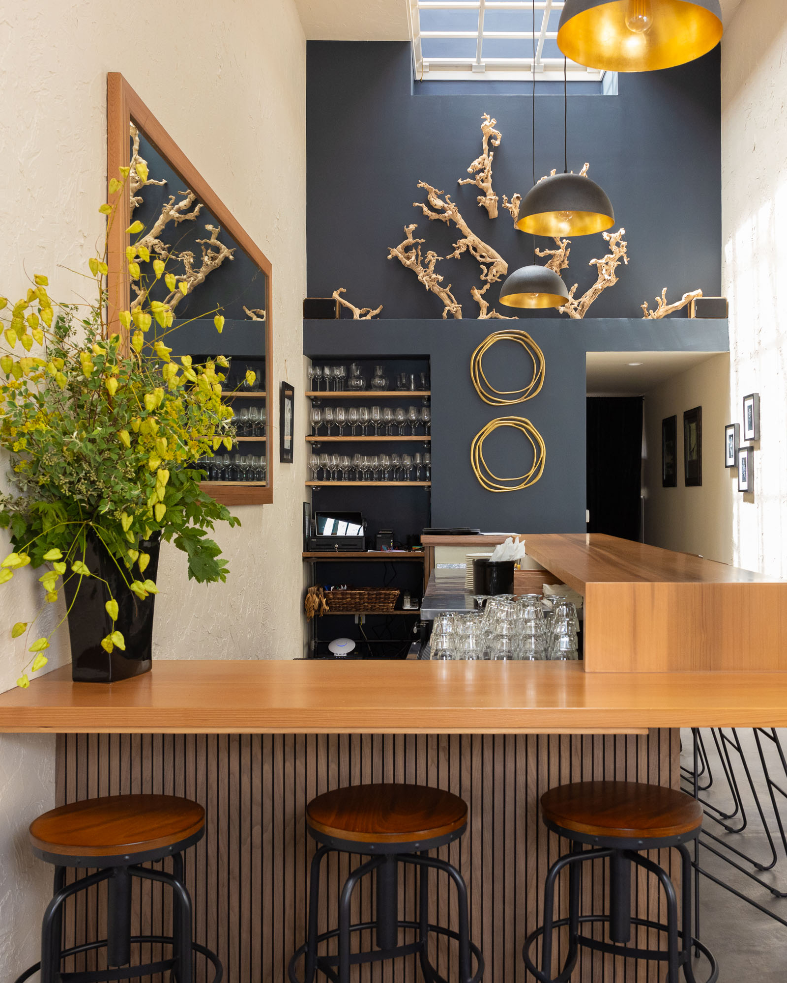 The image shows a modern bar with wooden stools and a counter. Behind are shelves with glasses, decorative wall art, plants, and large pendant lights.