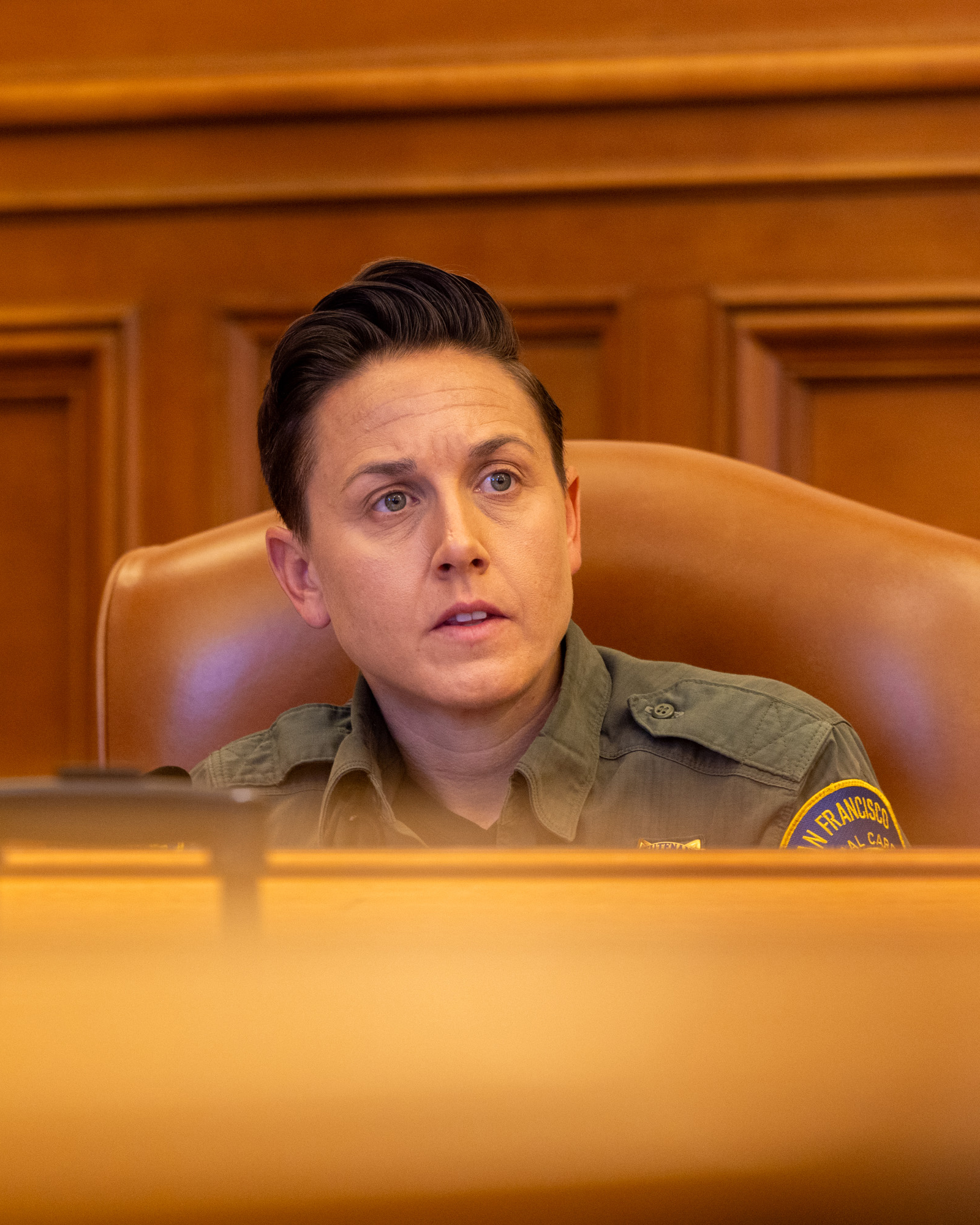 A person in a uniform sits in a brown leather chair behind a wooden desk, appearing engaged and attentive, with a polished wooden panel background.