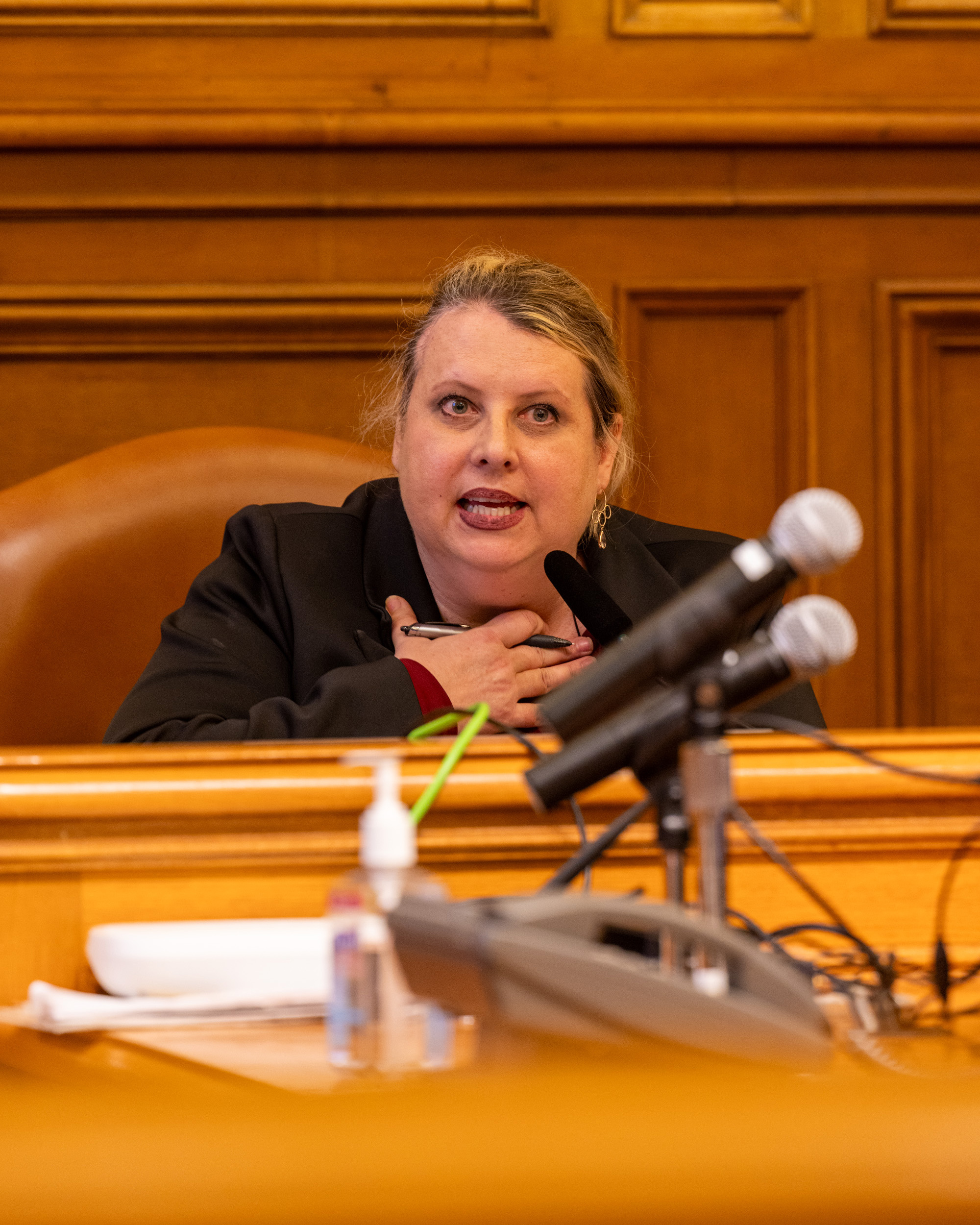 A woman sits at a wooden desk, speaking into a microphone. She appears to be in a formal setting, possibly a courtroom or meeting room, gesturing with her hand.