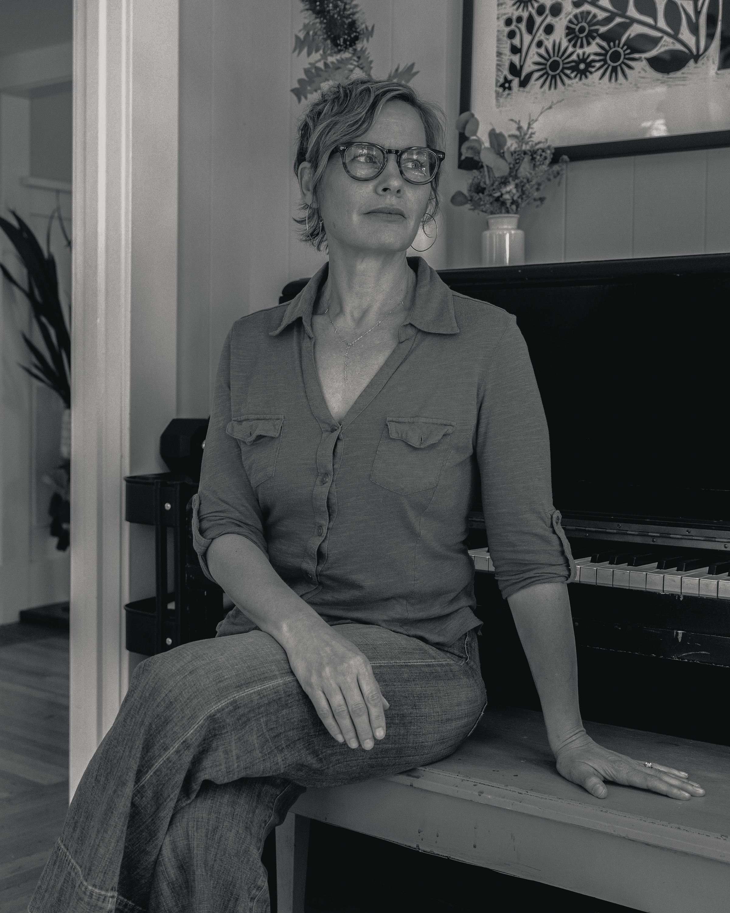 A woman with glasses and short hair sits on a bench, her right hand resting on a piano. She wears a buttoned shirt and jeans, set in a room with a framed artwork.