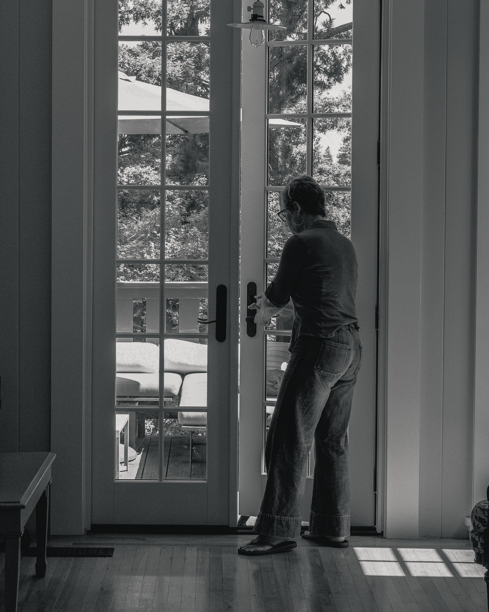 A person stands indoors, opening a French door looking toward an outdoor patio with furniture and trees in the background. The setting is serene and monochromatic.