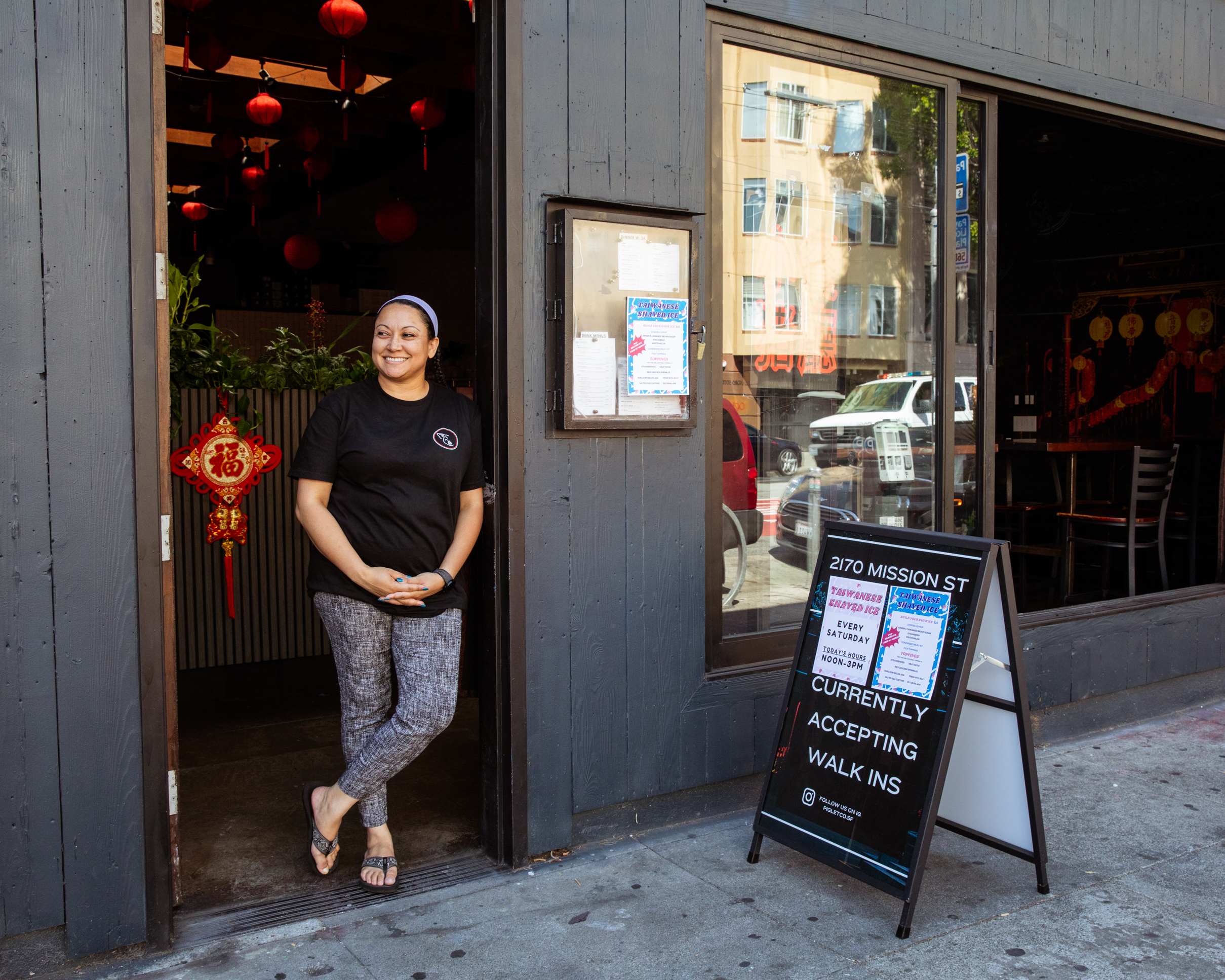 A smiling woman stands at a doorway, decorated with a red lantern and sign, at a dimly lit restaurant. Outside, a sidewalk sign invites walk-ins.