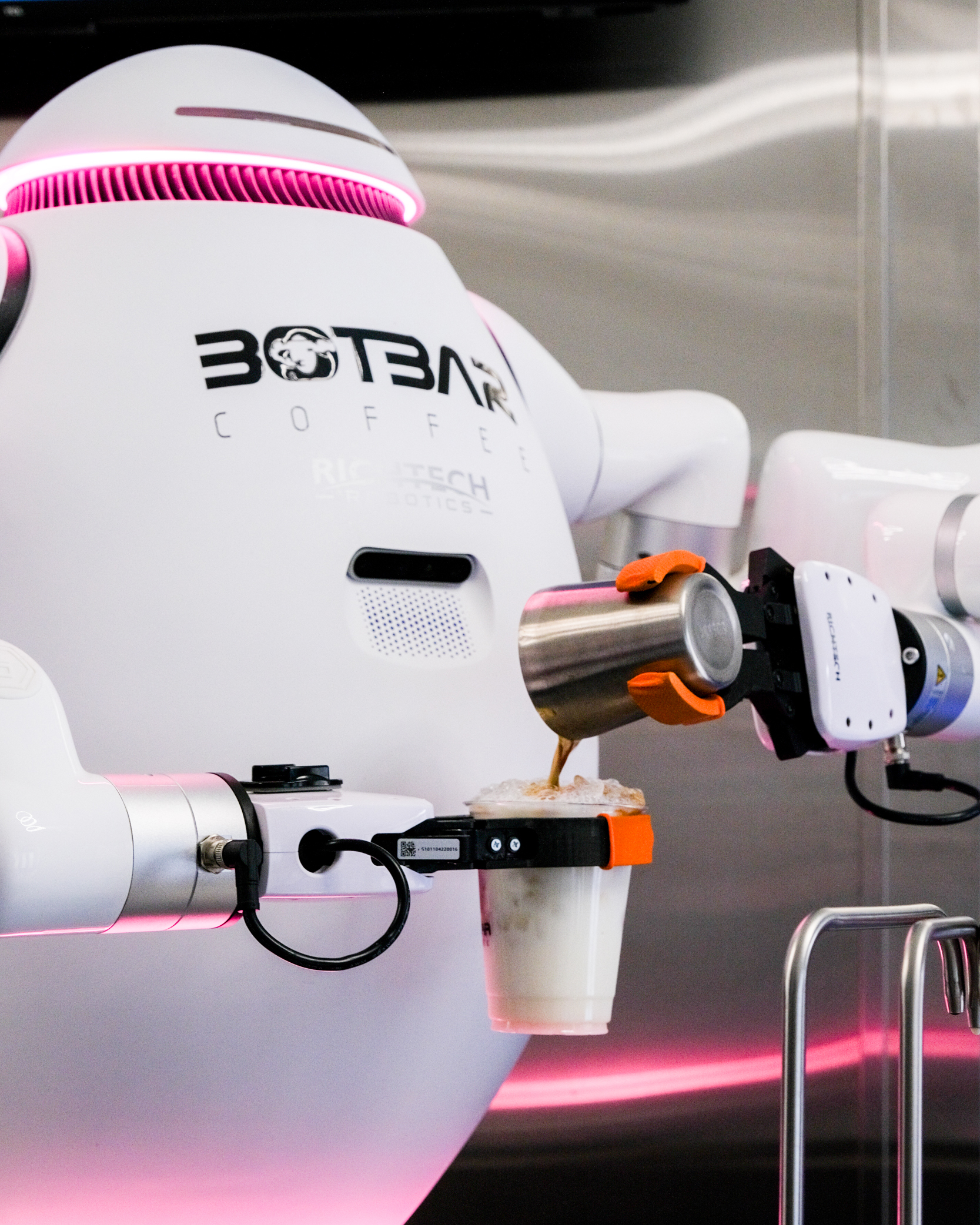 A white robot with &quot;Botbar Coffee&quot; on it pours a foamy beverage into a clear cup. Its robotic arm holds the cup while the other arm pours the drink.