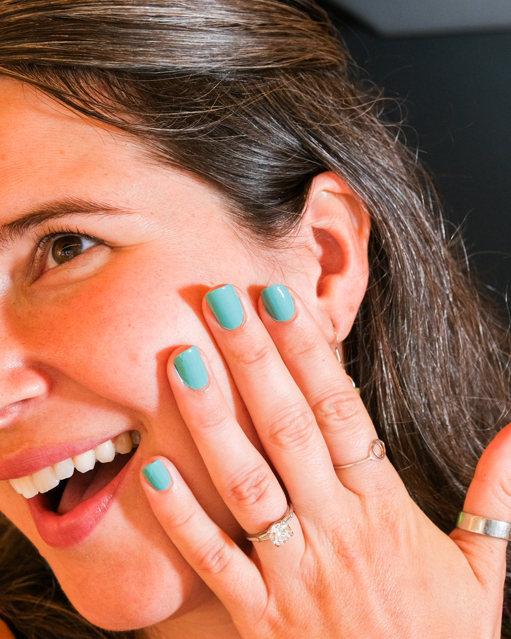 A person with long brown hair smiles widely, showcasing white teeth. The face is partially covered by a hand with teal nail polish and wearing several rings.