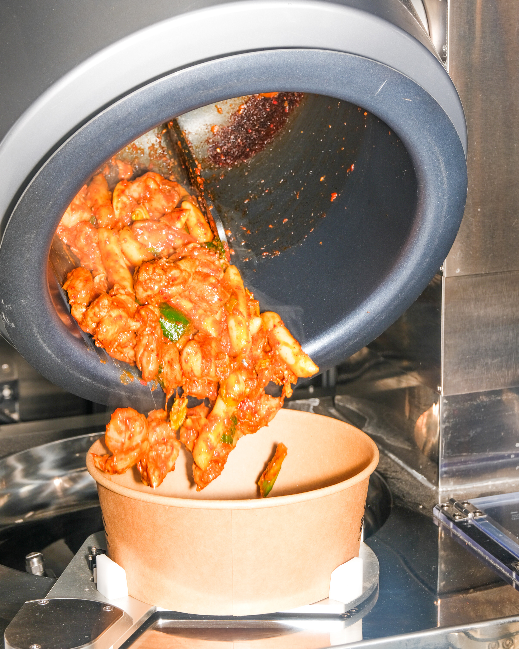 A robotic arm is pouring a spicy, saucy dish into a brown paper bowl from a large metal container. The dish appears to be a mixture of meat and vegetables.