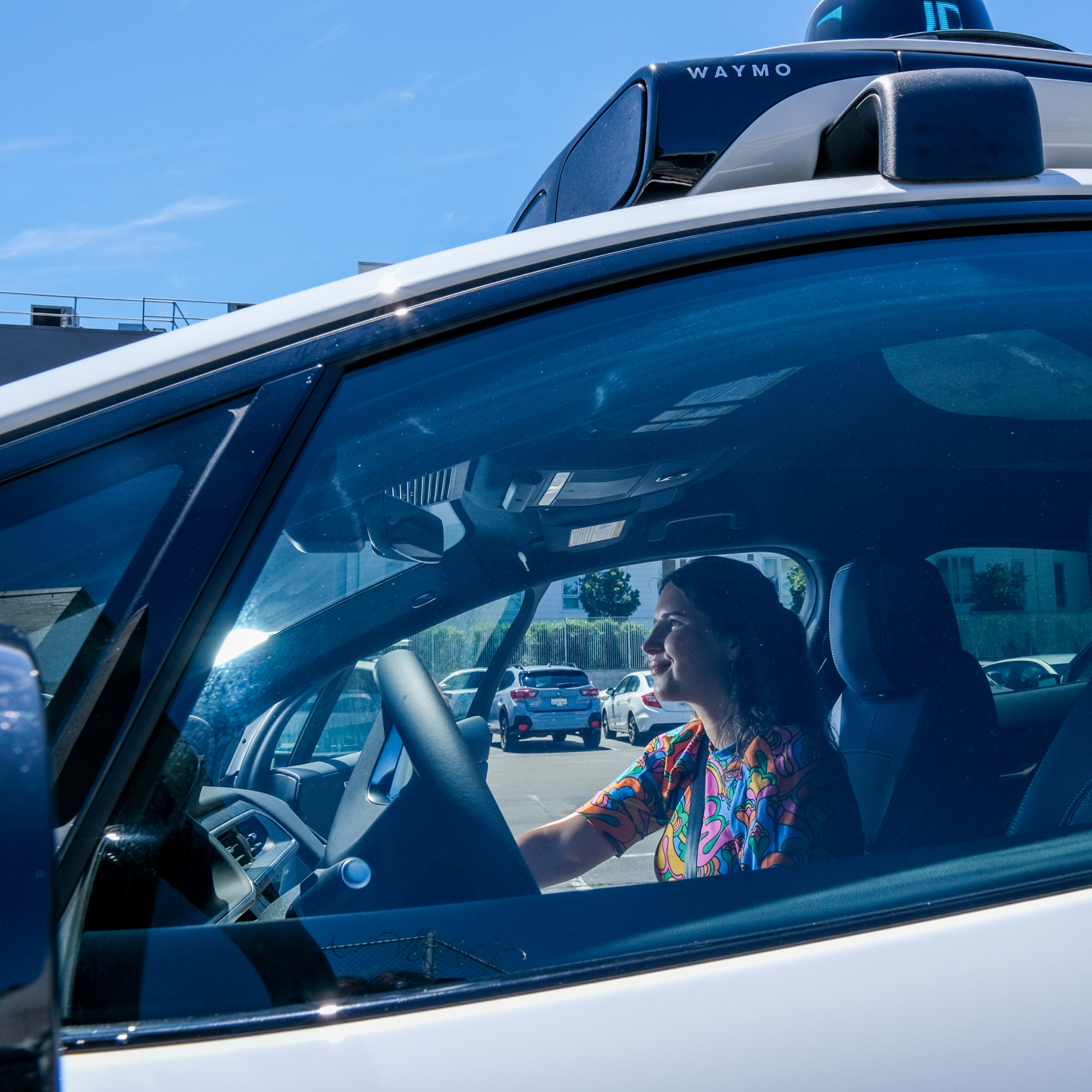 A woman is smiling while sitting in the driver's seat of a white car with a &quot;Waymo&quot; logo on top, indicating it's a self-driving vehicle.