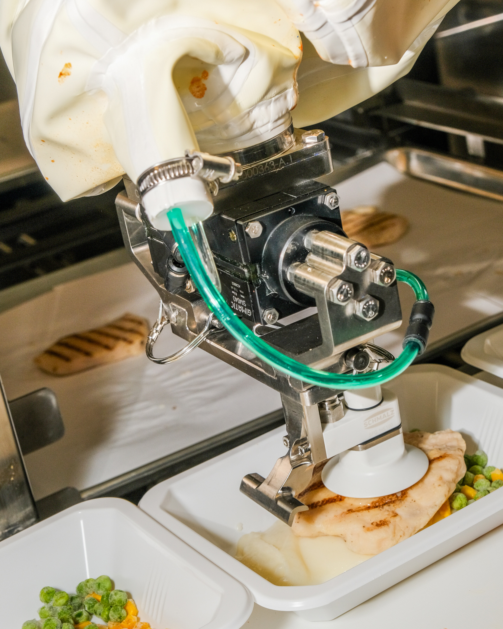 A robotic arm is placing a piece of grilled chicken on a plate with mashed potatoes and mixed vegetables in an industrial food preparation setting.