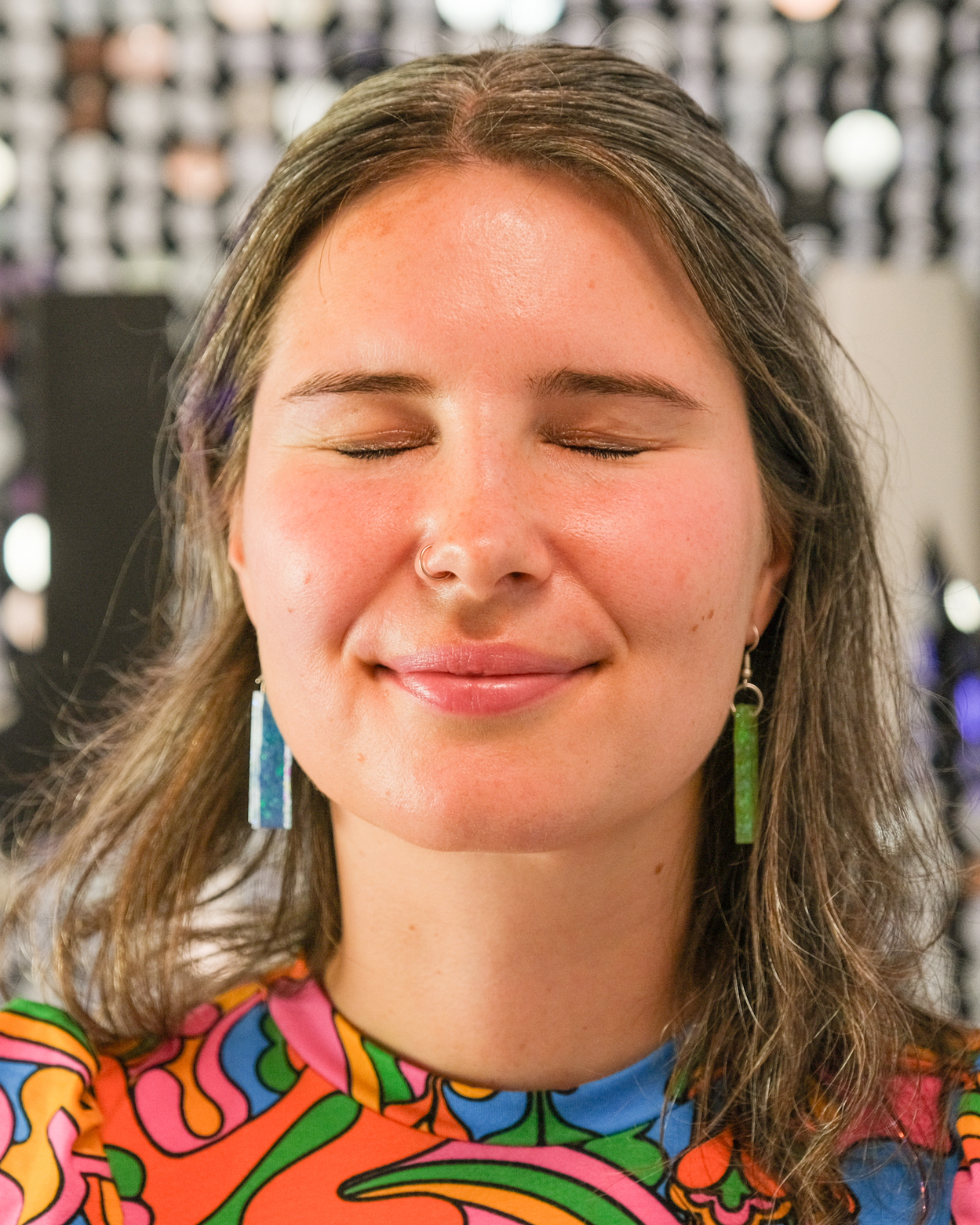 A woman with closed eyes and a slight smile is wearing colorful, patterned clothing and rectangular green-blue earrings. The background is blurred with circular lights.