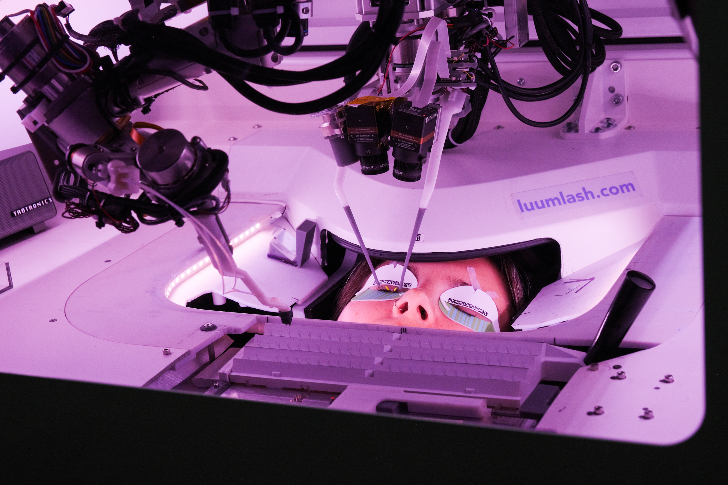 A person is lying under a machine with their eyes covered, surrounded by intricate robotic arms and equipment, marked with "luumlash.com" in an illuminated environment.