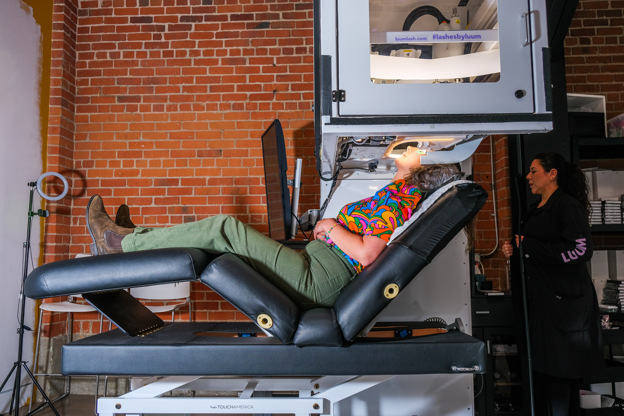 A person lies on a reclined chair with their head under a machine, while another person stands nearby in a room with exposed brick walls.