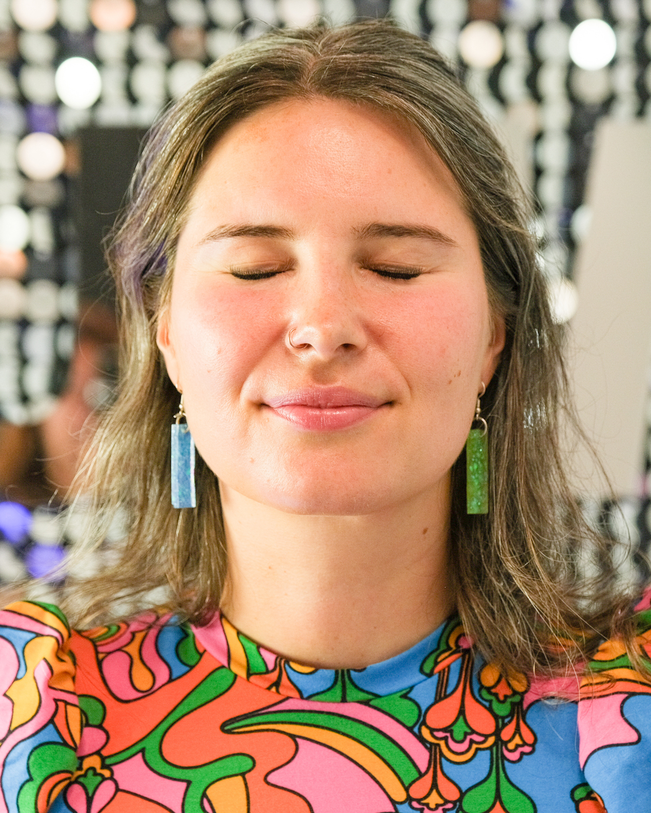 A woman with long hair and closed eyes wears colorful, patterned clothing and sparkly green earrings, standing against a blurred background of circular lights.