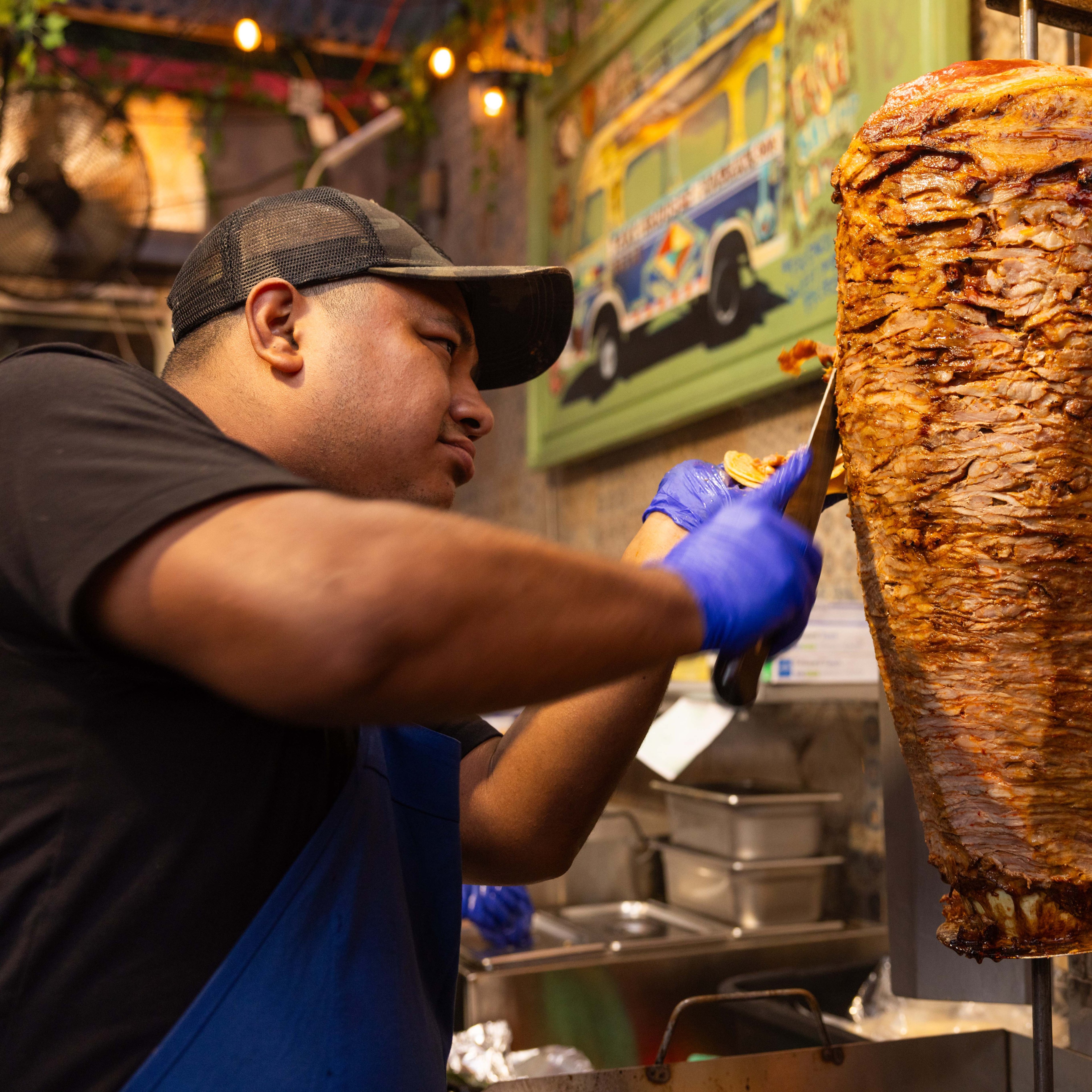 A man in a blue apron and cap carves meat from a vertical rotisserie in a kitchen, with a colorful bus painting on the wall behind him.