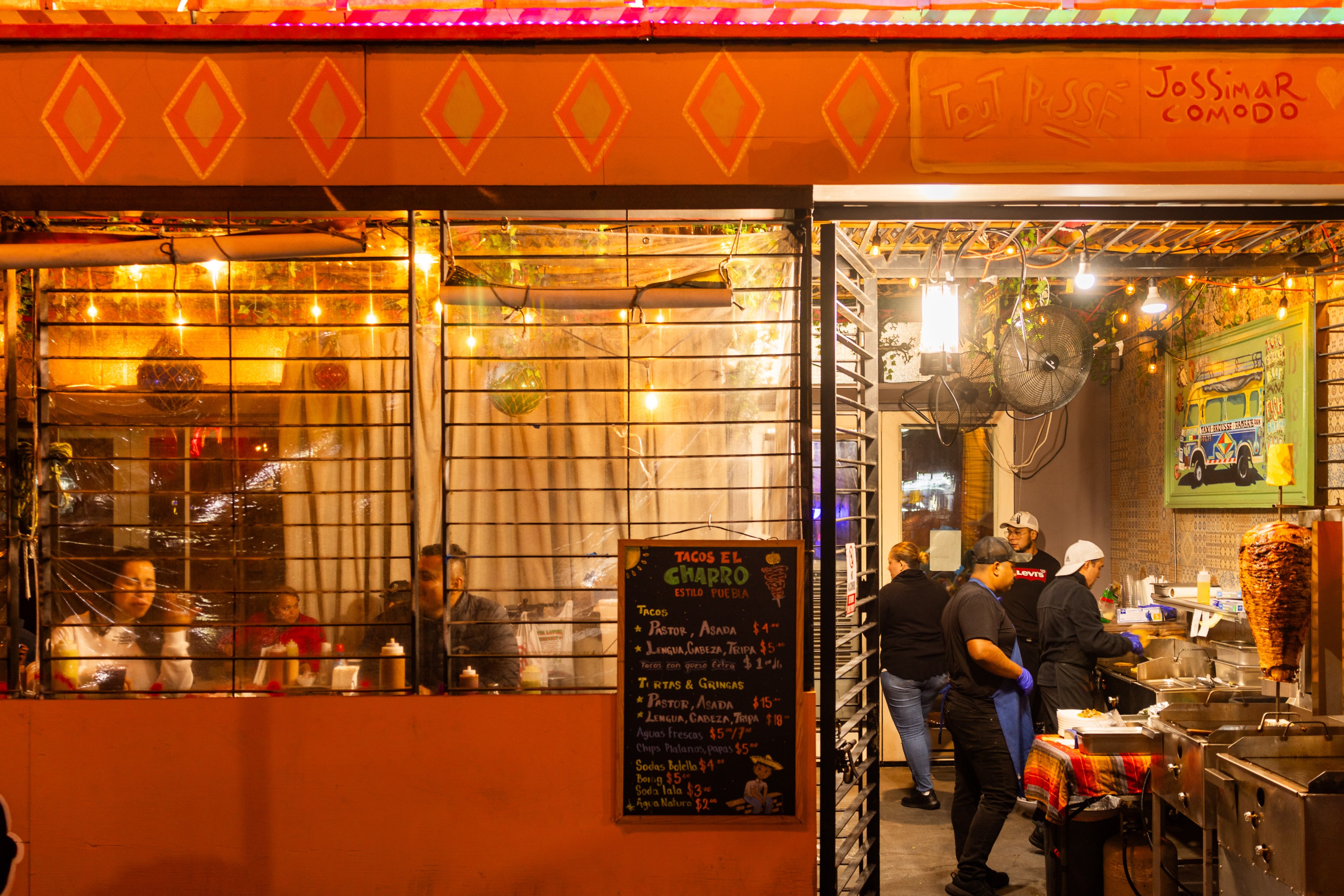 A lively, brightly lit taco stand has patrons dining on the left and cooks preparing food on the right. A colorful menu board displays various taco options.