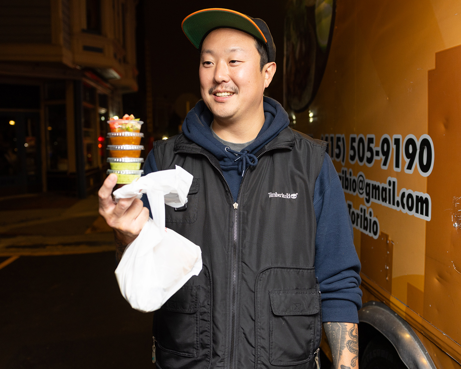 A smiling man in a black vest and green cap holds a stack of colorful containers and a white plastic bag, standing in front of an orange food truck at night.