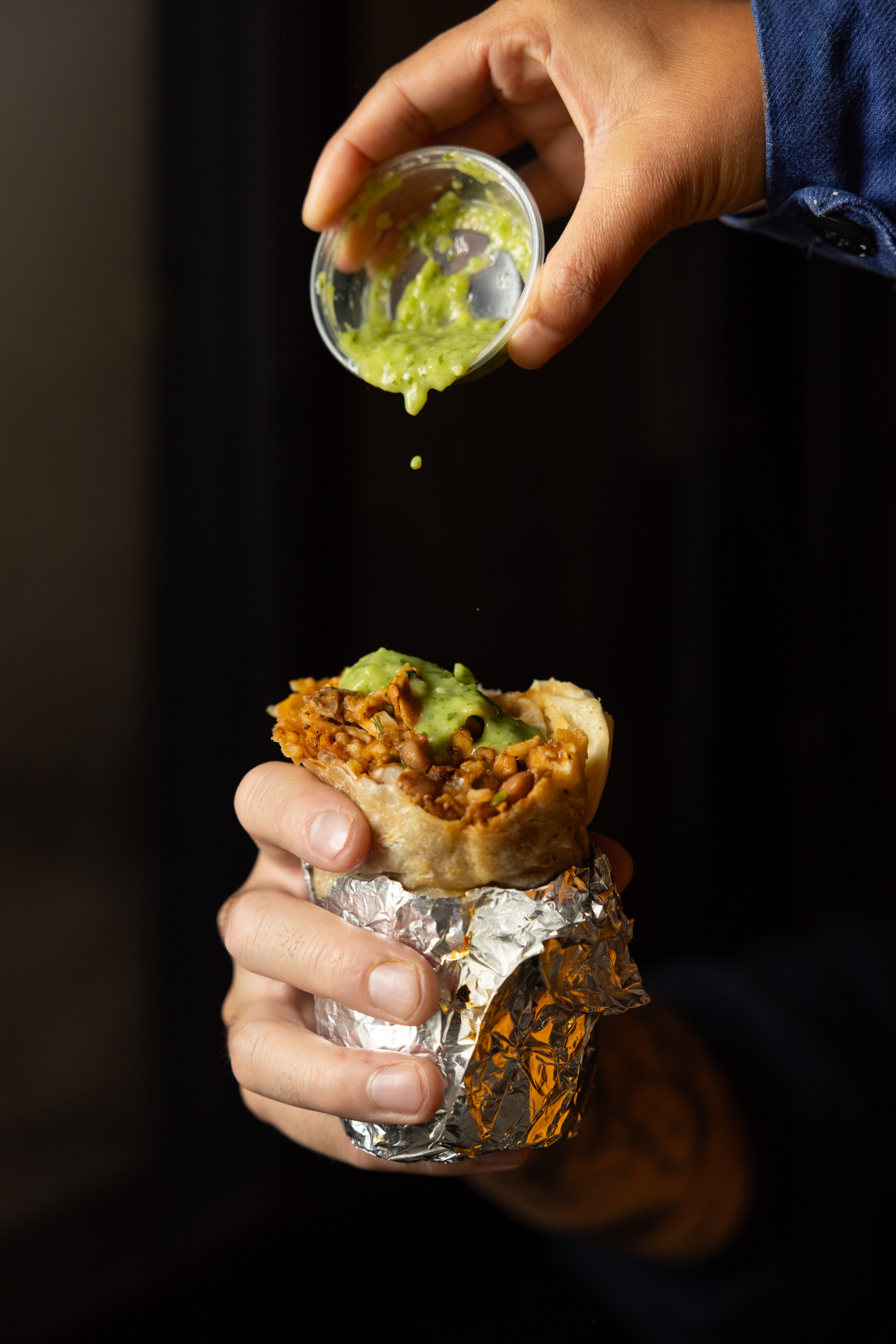 A hand holds a foil-wrapped burrito while another hand drizzles green sauce from a small cup onto it. The burrito is filled with rice, beans, and meat.