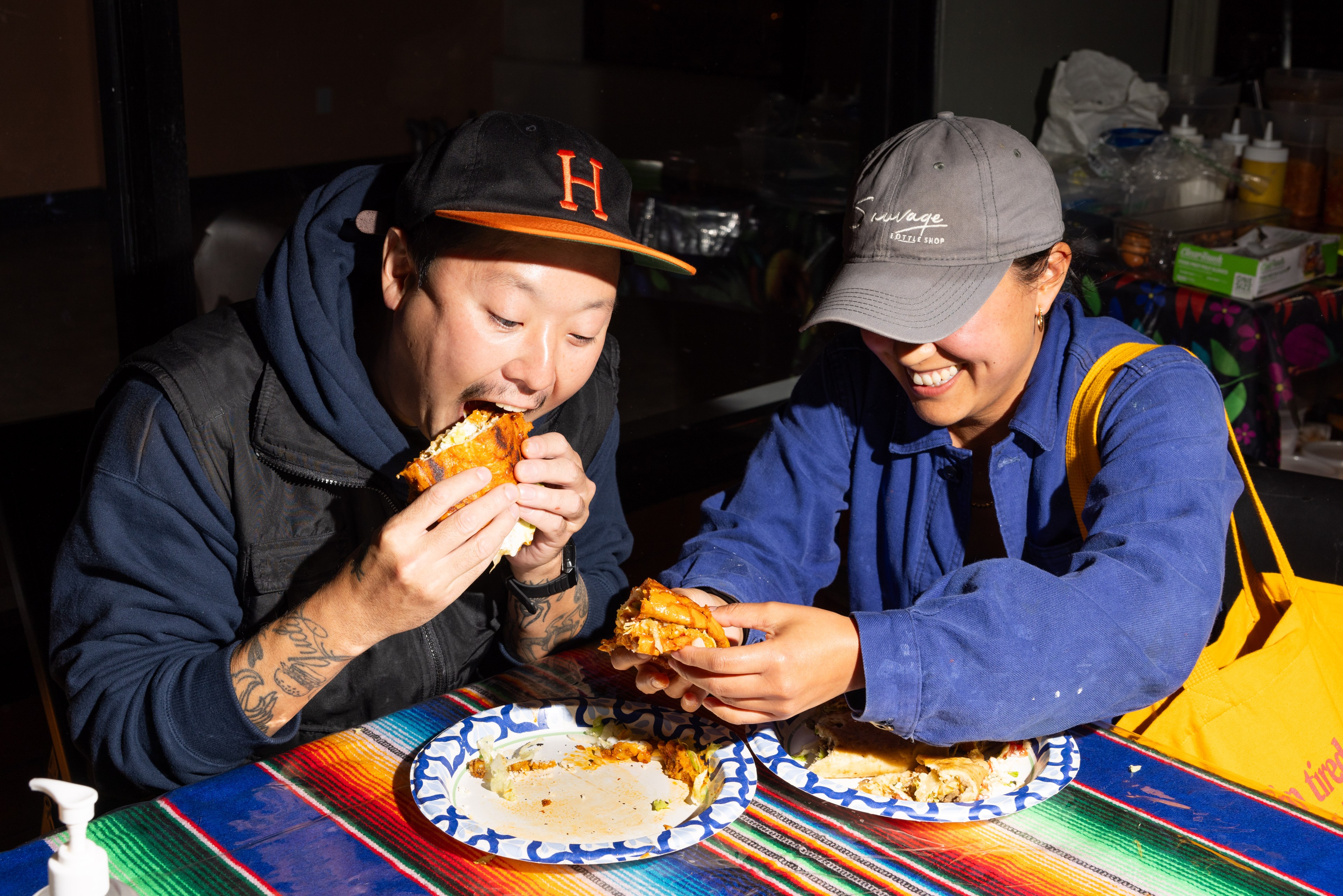 Two people are enjoying large sandwiches at a brightly colored table. They are both smiling and appear to be savoring their food, with paper plates in front of them.