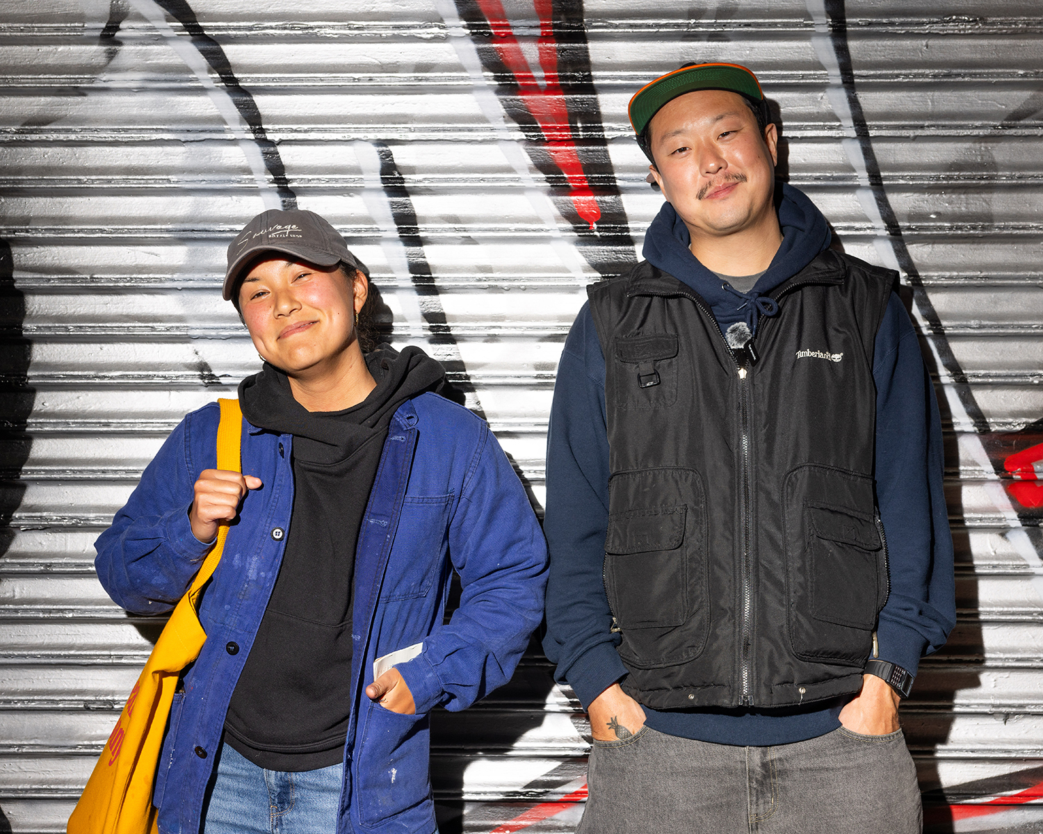 Two people stand in front of a graffiti-covered metal door, smiling. They wear casual clothes: jackets, hoodies, and caps, one holding a yellow bag.