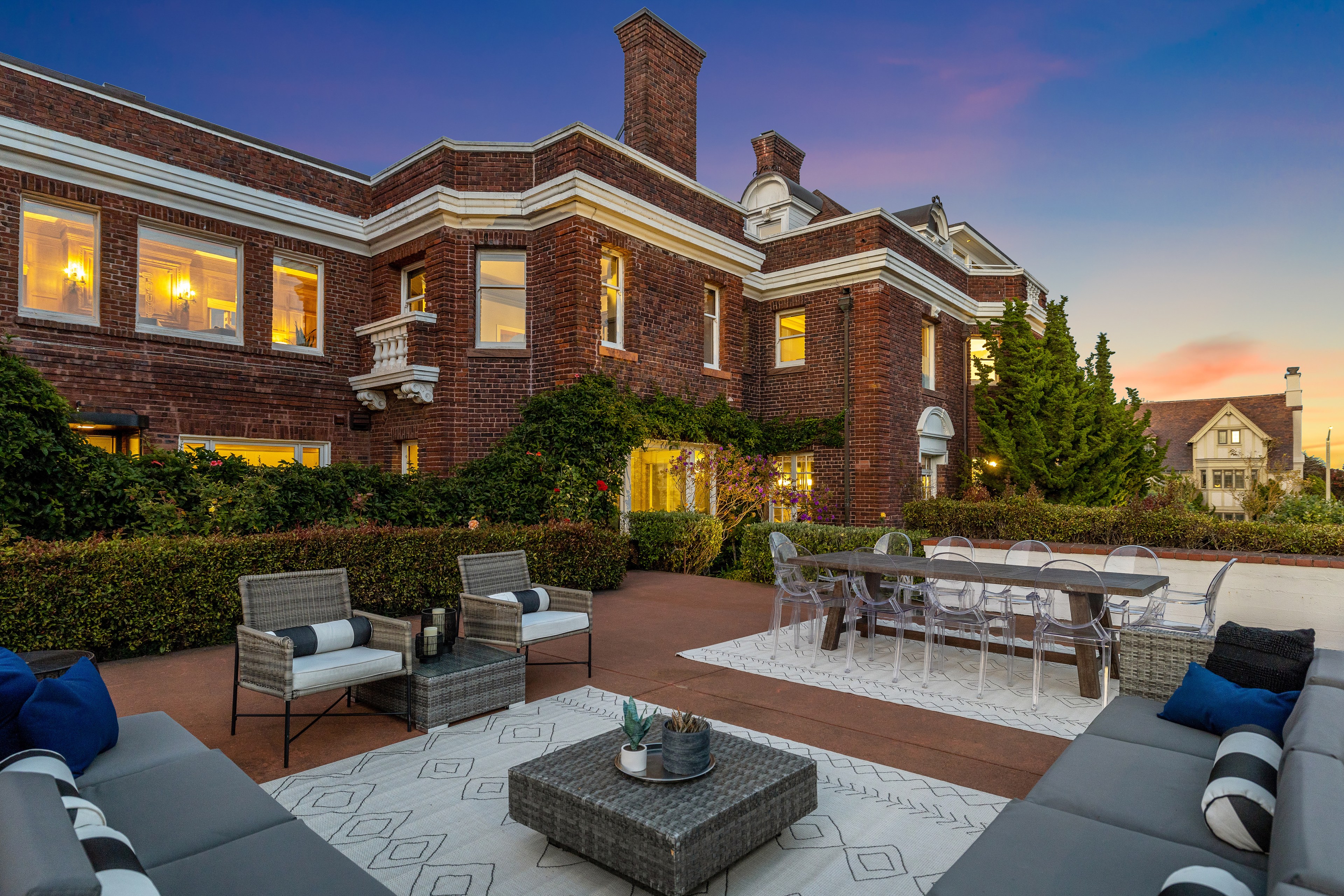 The image shows a beautiful brick building with lush landscaping and a spacious outdoor patio. The patio has modern furniture, including sofas, chairs, and a dining table.