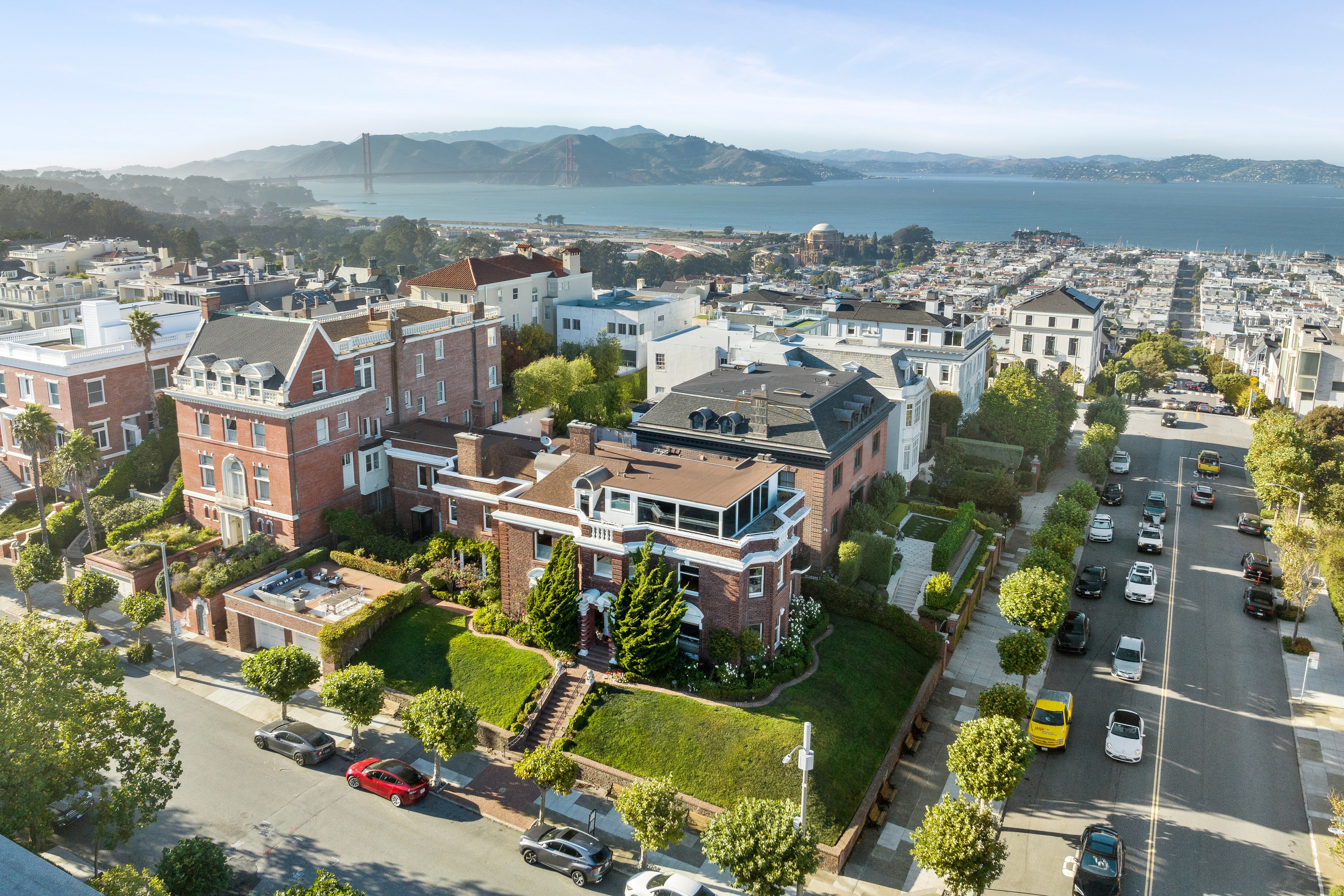 The image shows a scenic neighborhood with elegant brick houses, lush gardens, and tree-lined streets, featuring a backdrop of mountains and a bay with a prominent bridge.