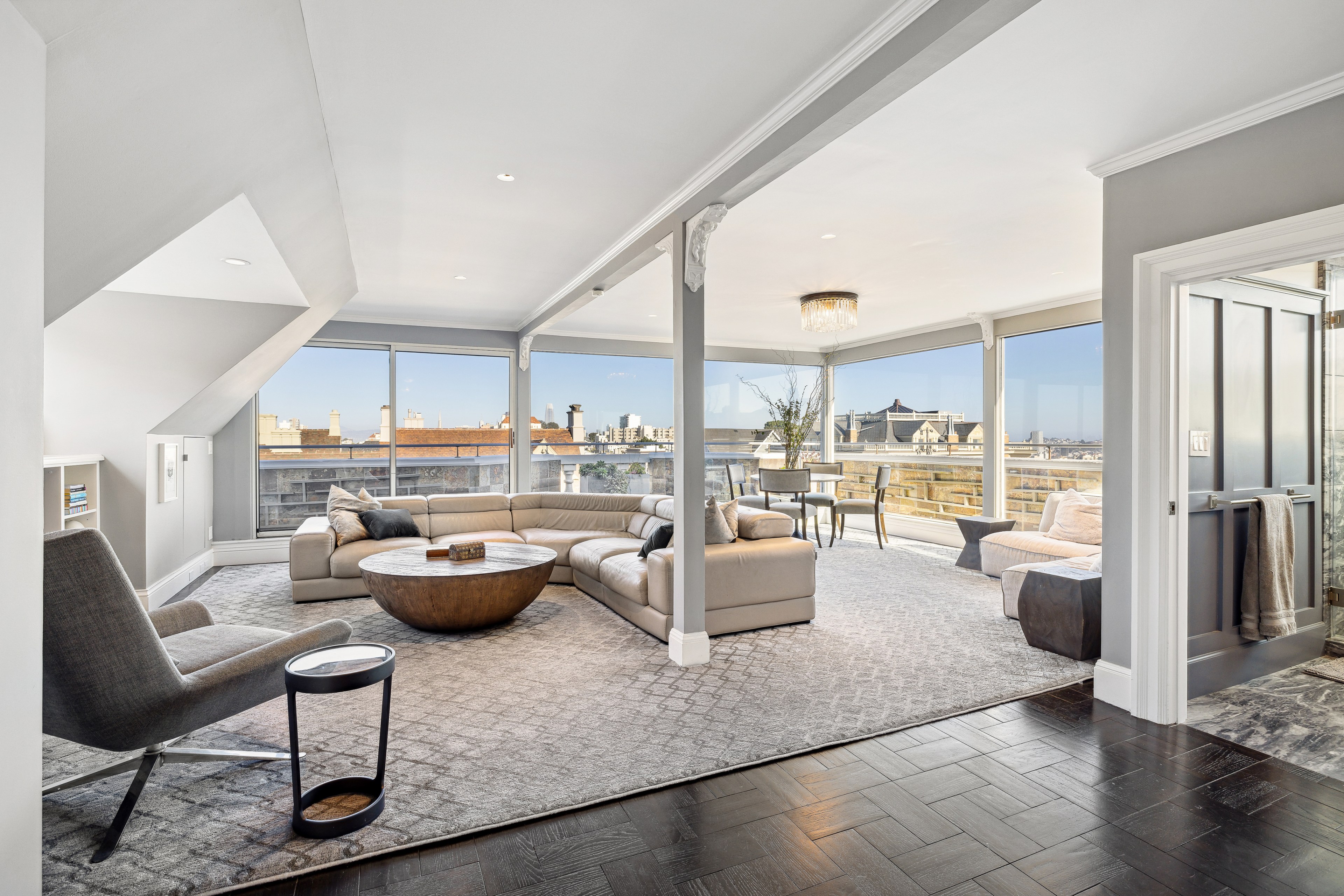 A spacious living room with gray walls and carpet features a sectional sofa, round coffee table, and modern armchairs. Large windows offer a city view and natural light.