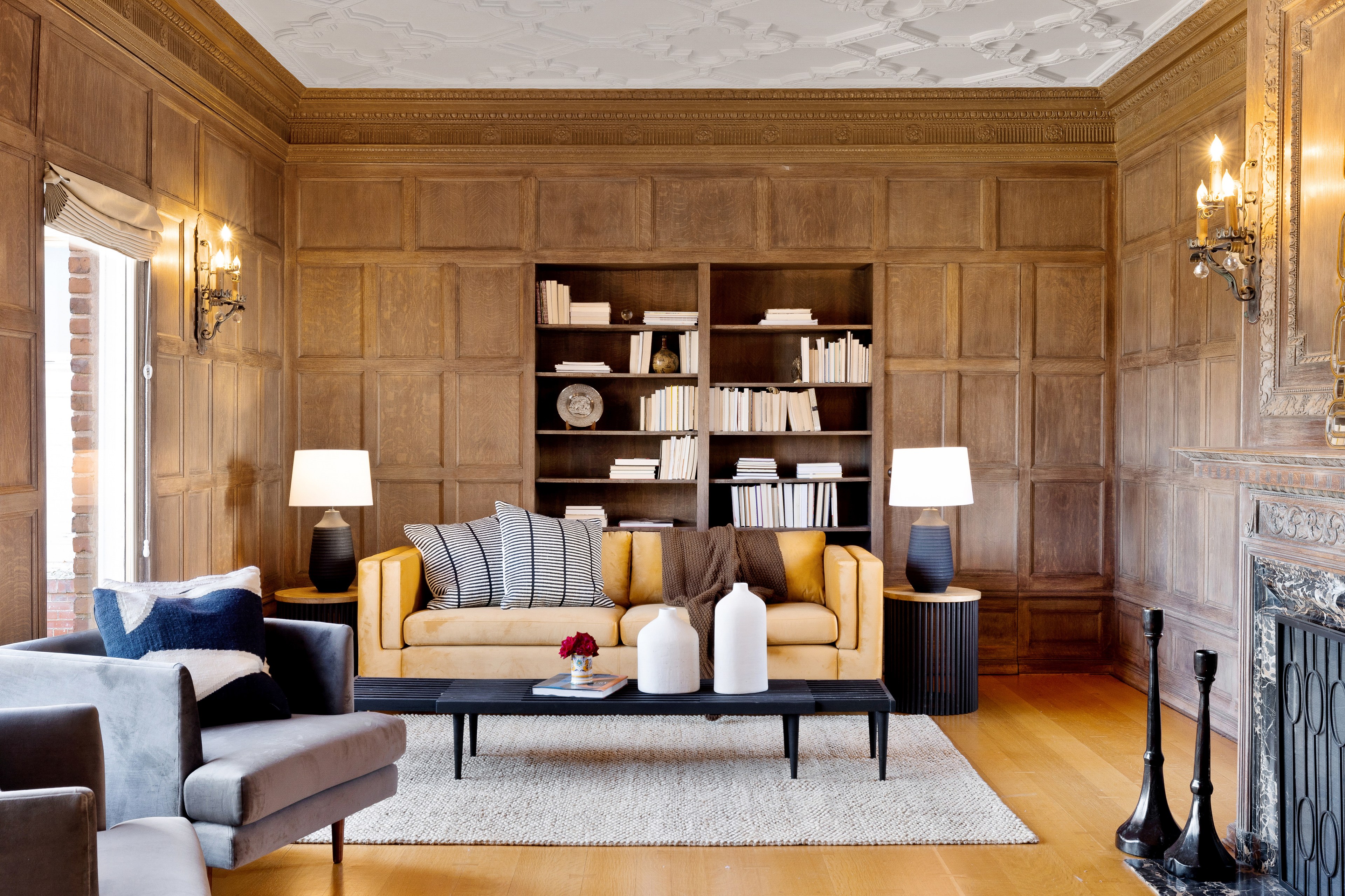 A cozy living room with wooden paneled walls, a yellow sofa with striped pillows, grey armchairs, a black coffee table, and bookshelves in the back.