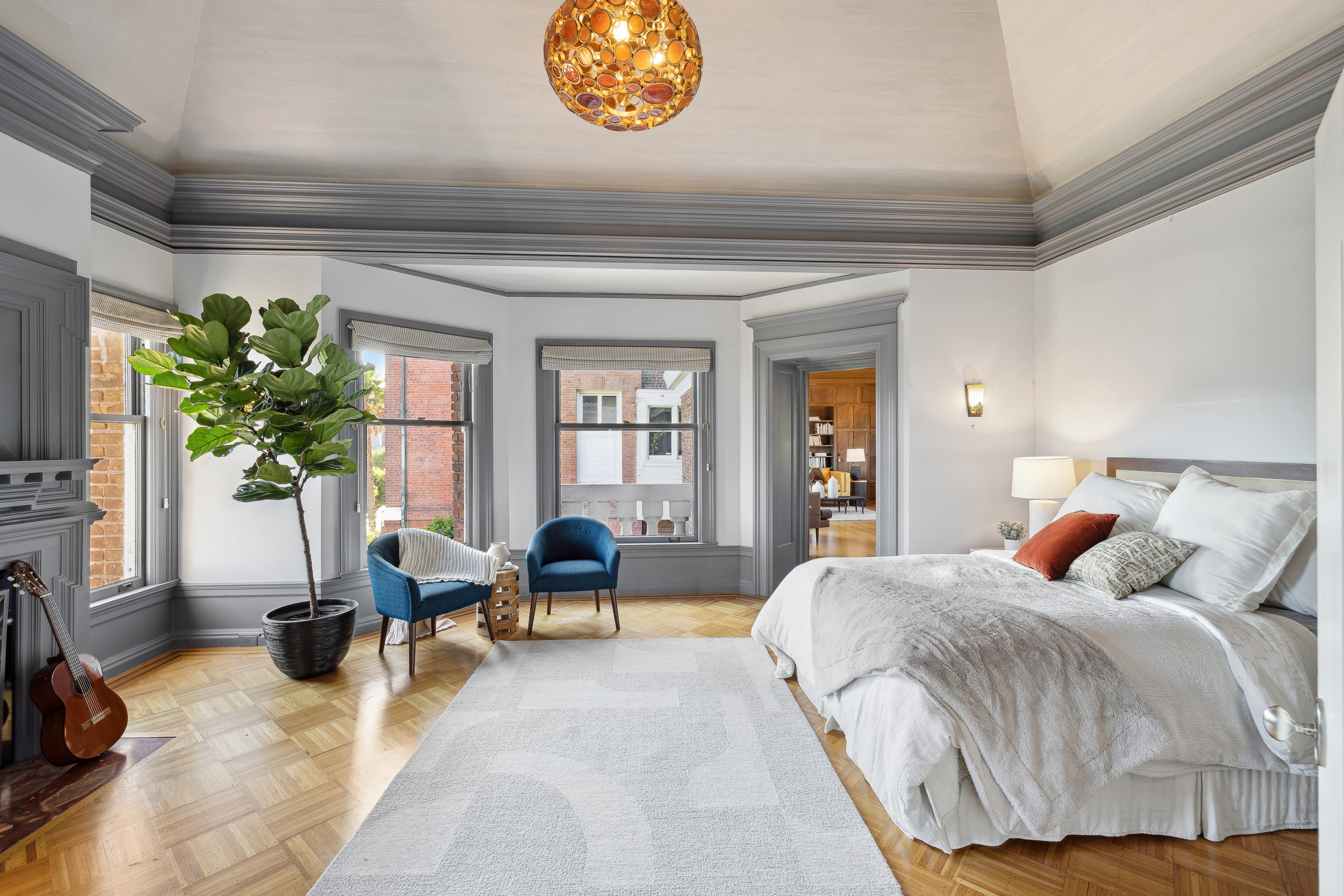 A cozy bedroom features a large bed with textured blankets, a potted plant, a guitar, two blue chairs, windows with a brick view, and a colorful hanging light.