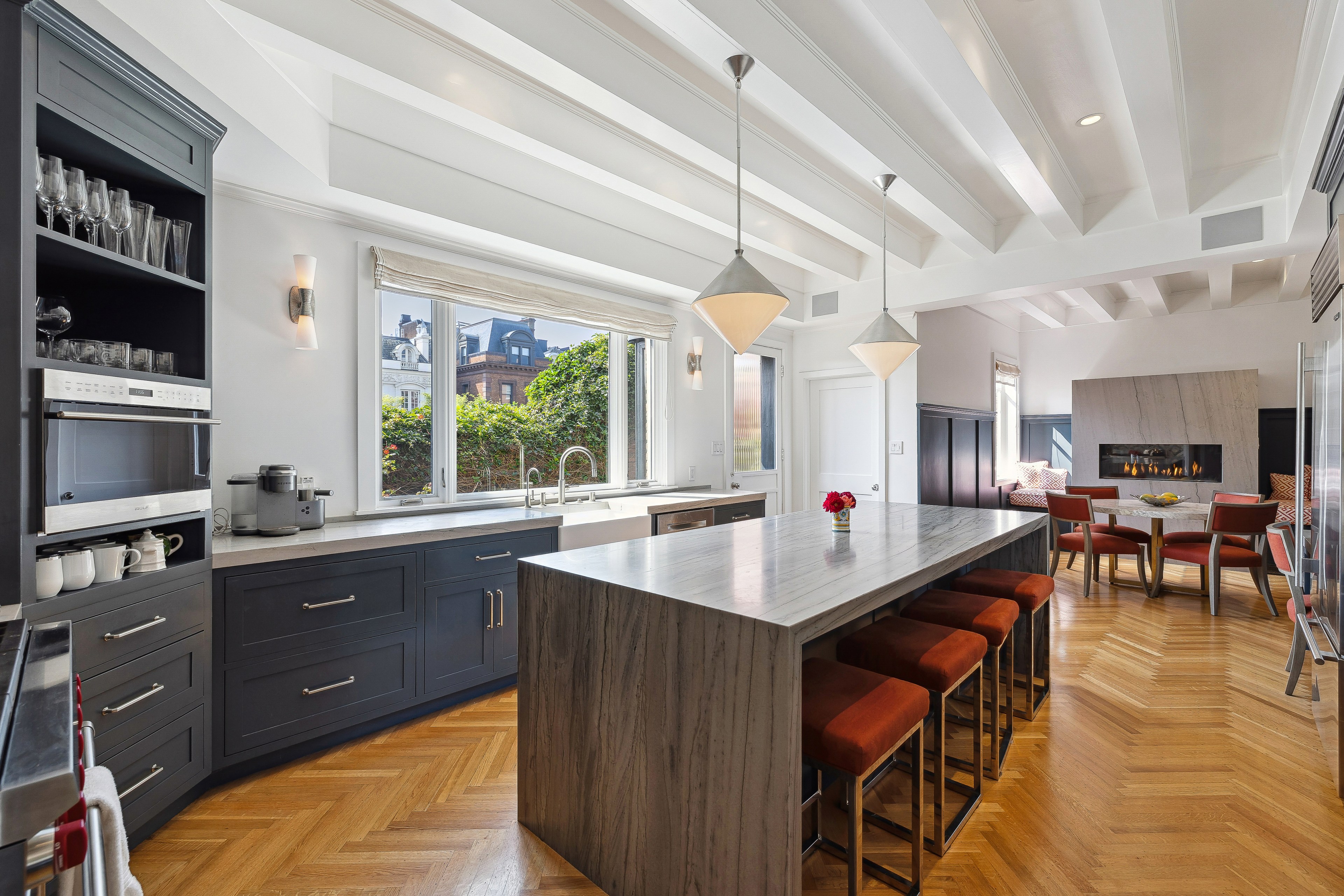 The image shows a modern kitchen and dining area with a large island, dark cabinetry, open shelving, and large windows. The dining area includes a table by a fireplace.