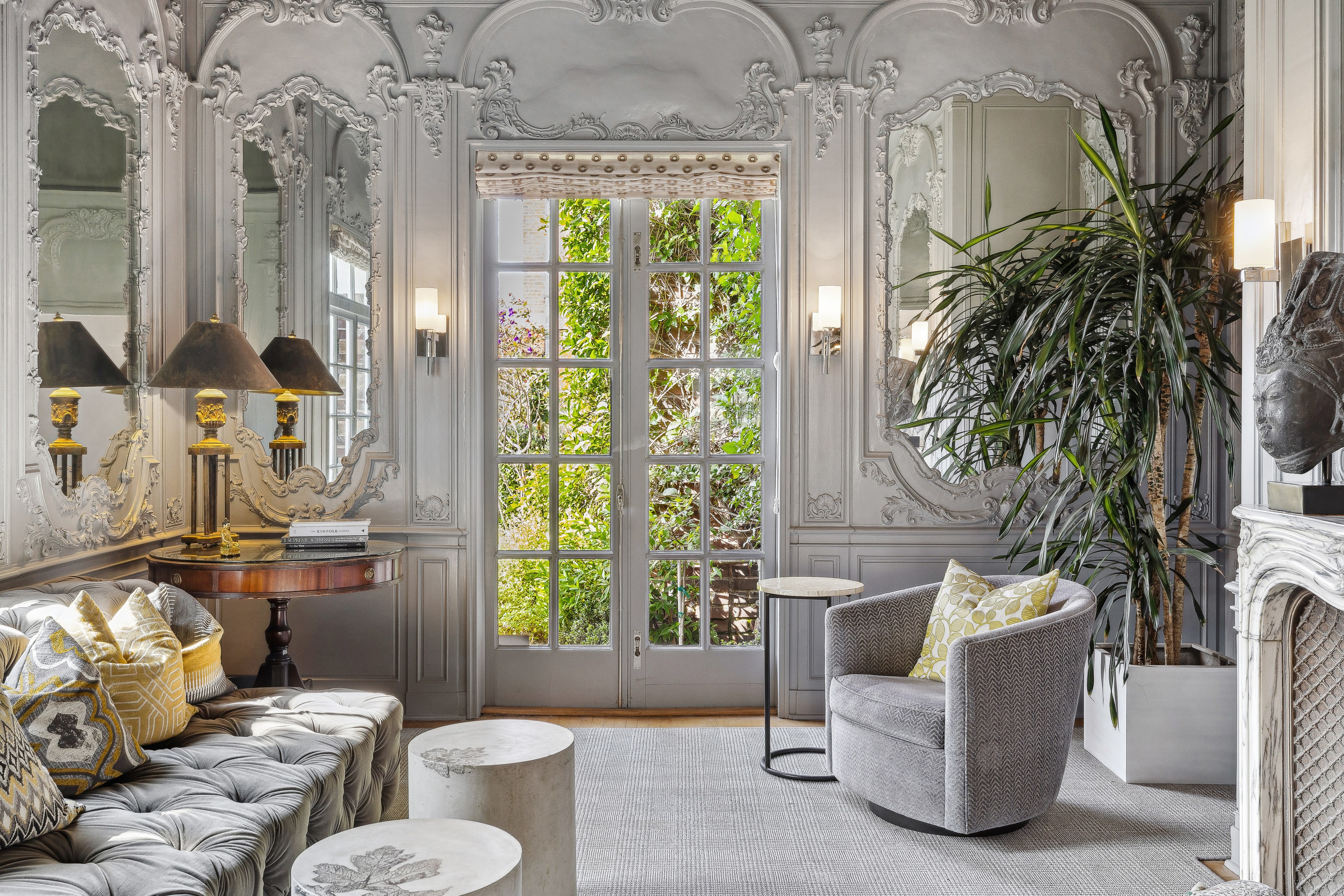 An elegant room features ornate mirrors, a tufted sofa, a round table with lamps, a plush chair, a tall plant, and French doors leading to a lush garden outside.