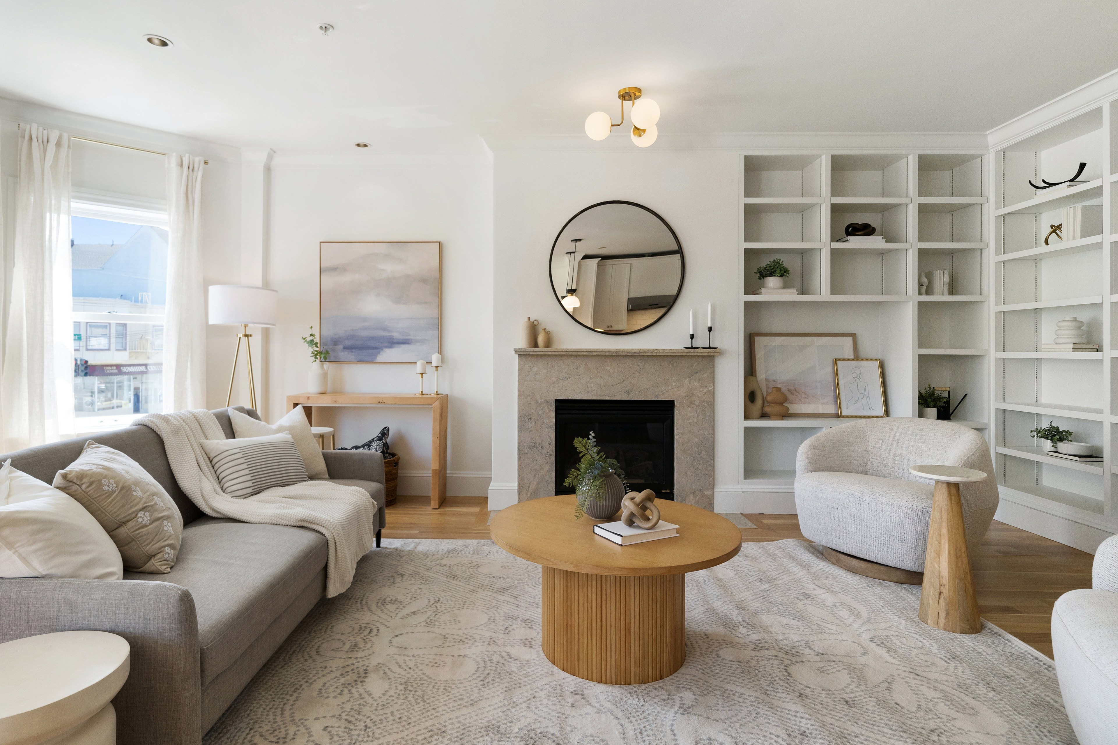 The image shows a modern living room with a light-colored sofa, wooden coffee table, round mirror above a fireplace, and white shelves with minimal decor.