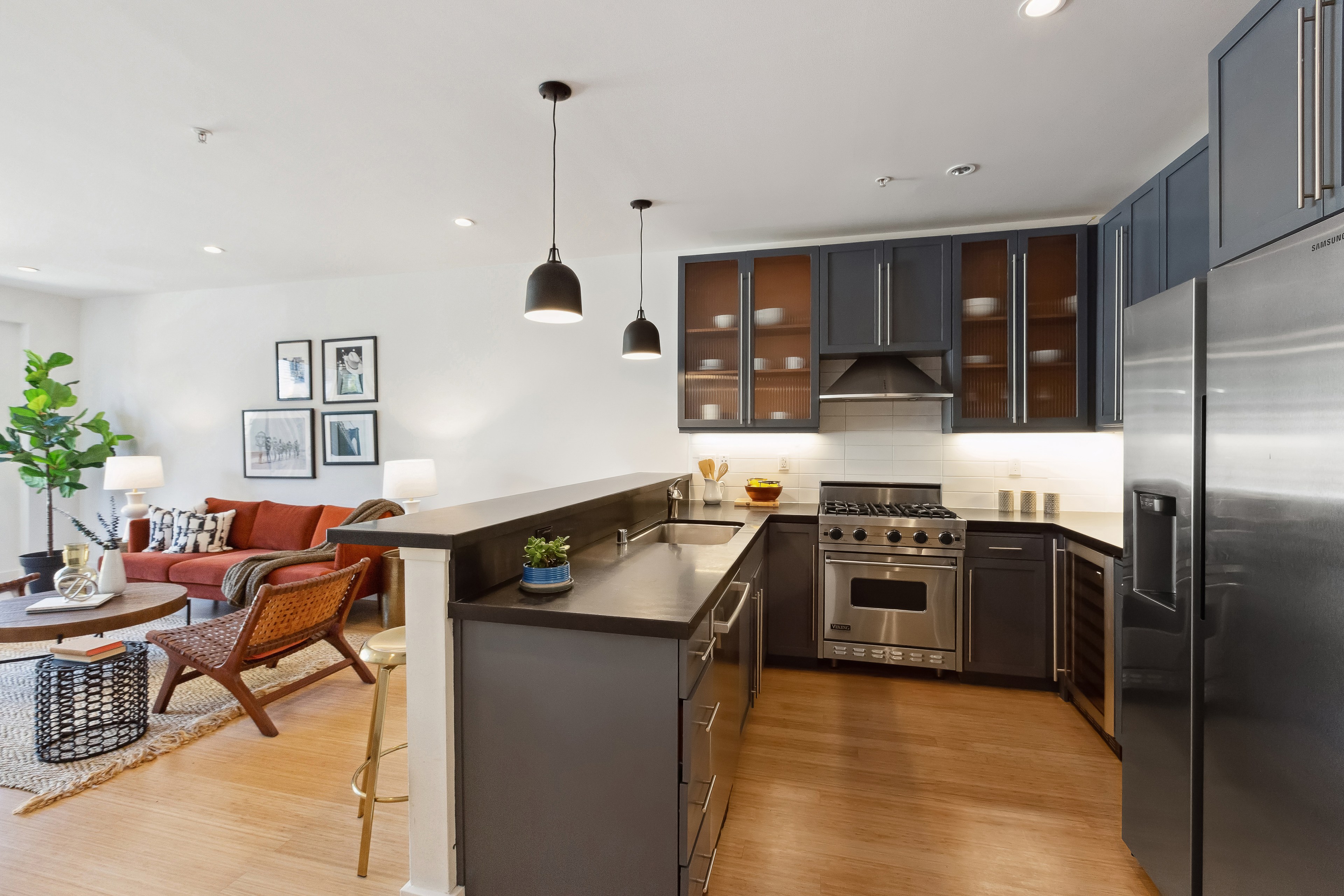 A modern kitchen with stainless steel appliances, dark cabinets, a central island with bar seating, and hanging pendant lights, open to a stylish living room with a red sofa.