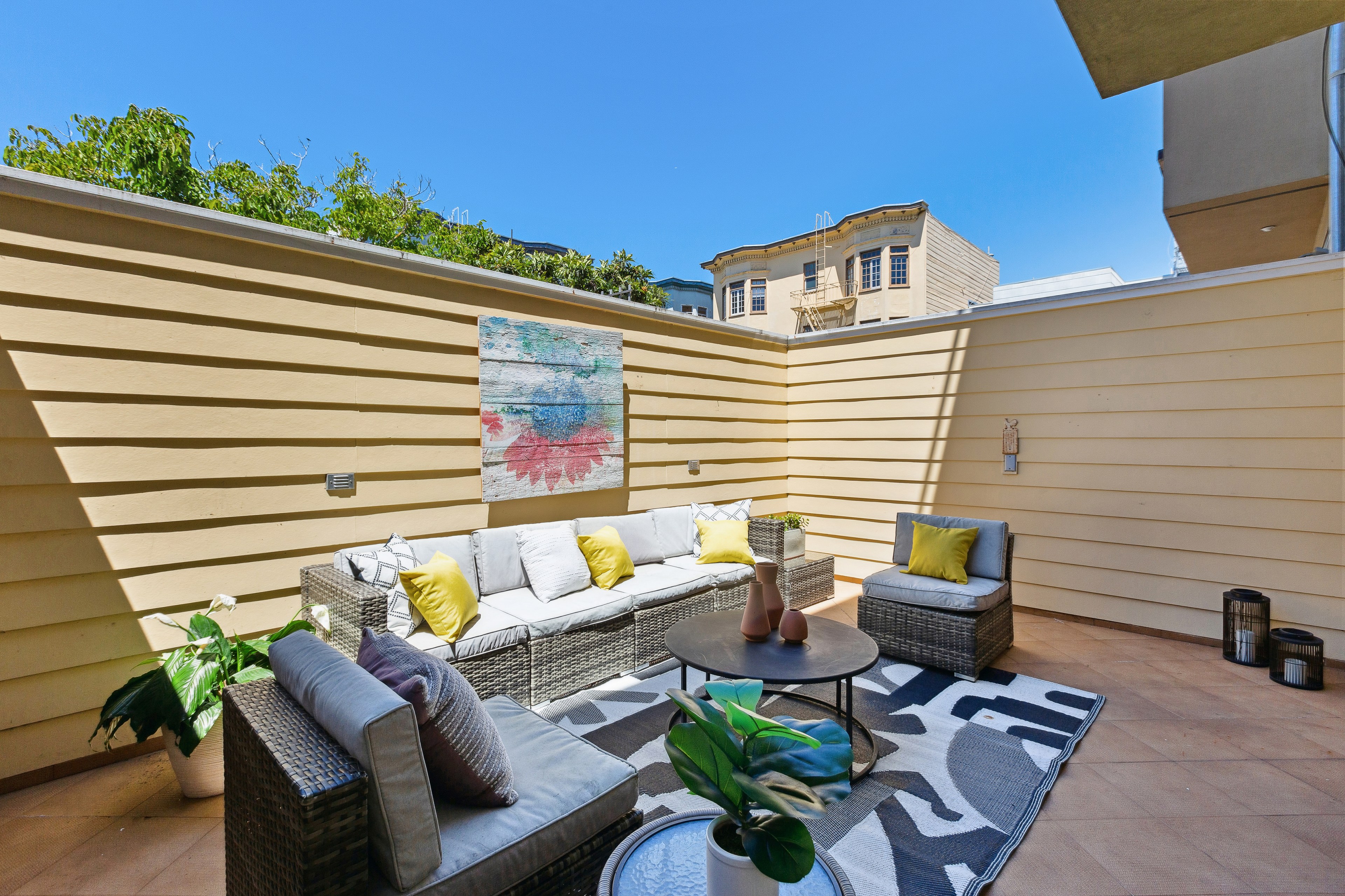 A cozy patio featuring a beige sectional sofa with yellow and white cushions, a round table with vases, and potted plants, all set against beige wooden walls with vibrant artwork.