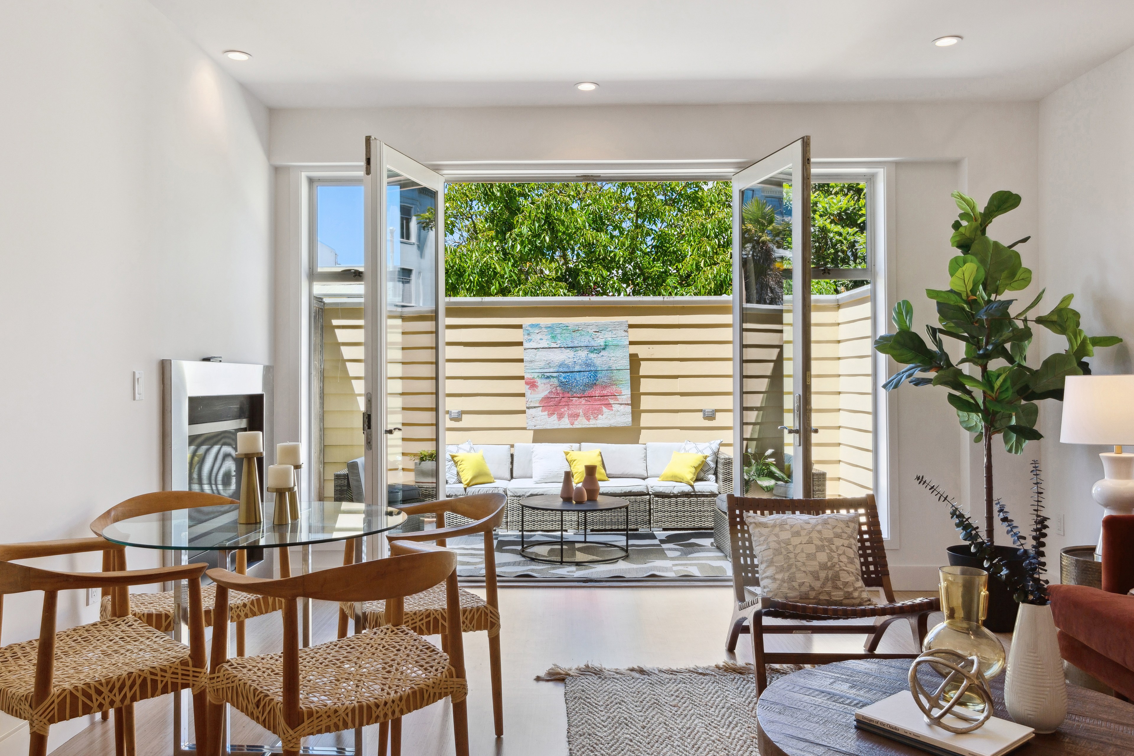 This image shows a bright, modern living space featuring a glass table with wooden chairs, an open patio door, and a cozy outdoor seating area with colorful cushions.