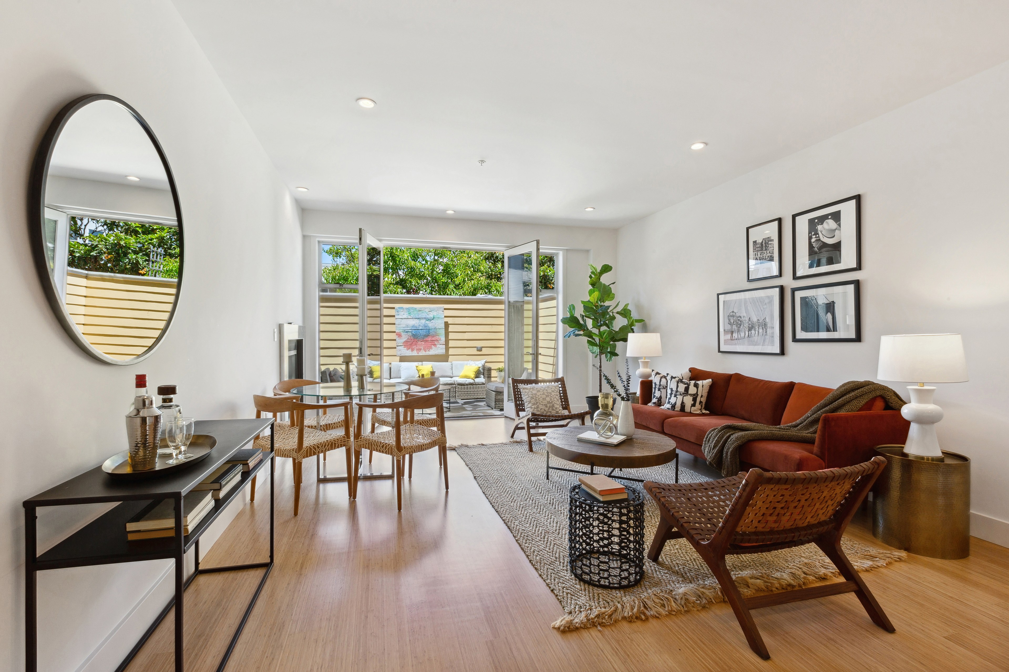 A modern living room with a red sofa, wooden chairs, and a glass coffee table. A large round mirror hangs on the wall, and glass doors open to an outdoor patio.