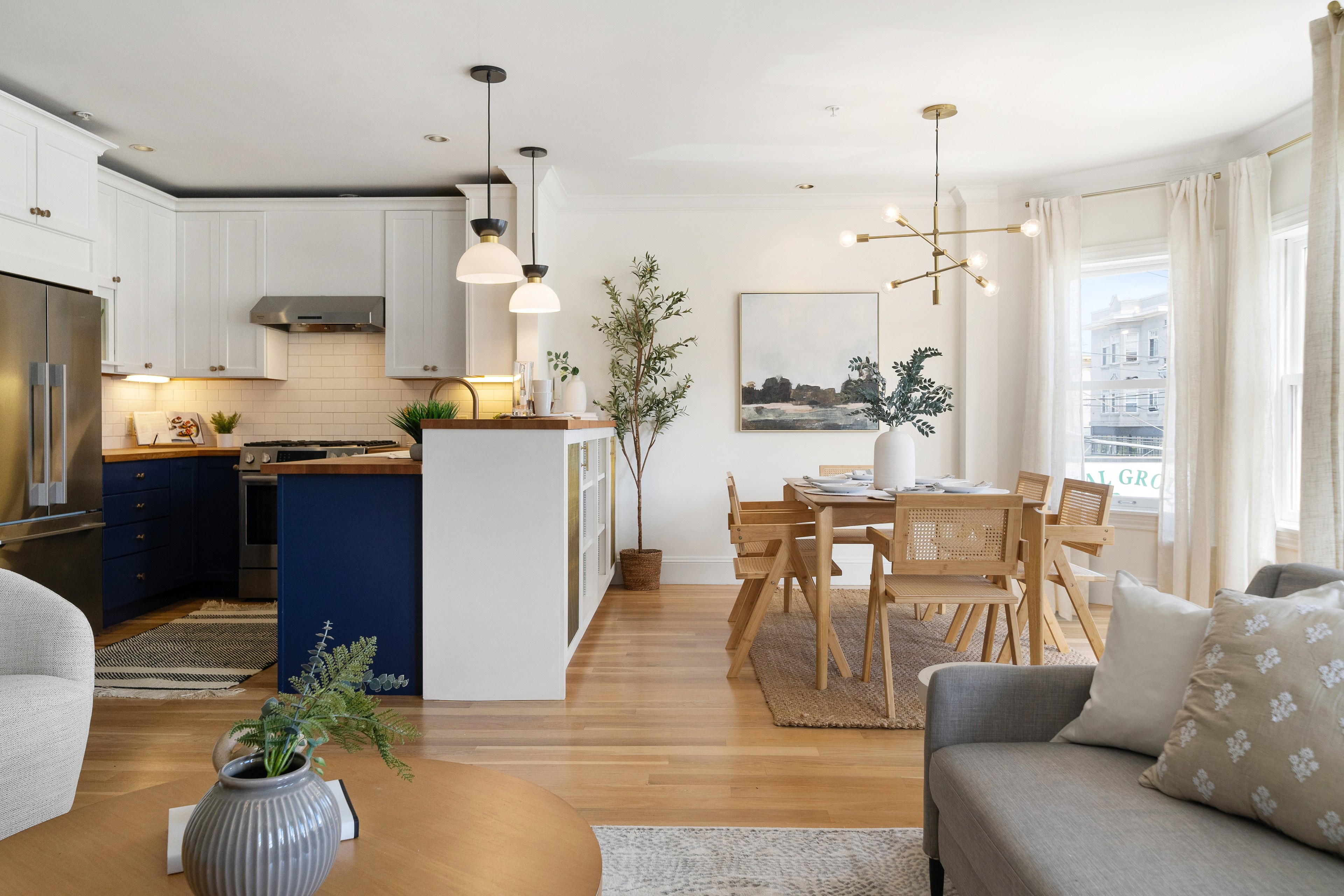 The image shows a modern open-concept kitchen and dining area. The kitchen has white cabinets, blue lower cabinets, stainless appliances, and wooden countertops. The dining area features wooden furniture, a modern chandelier, and wall art above the table. The living area has a gray sofa and a coffee table with a vase on it.