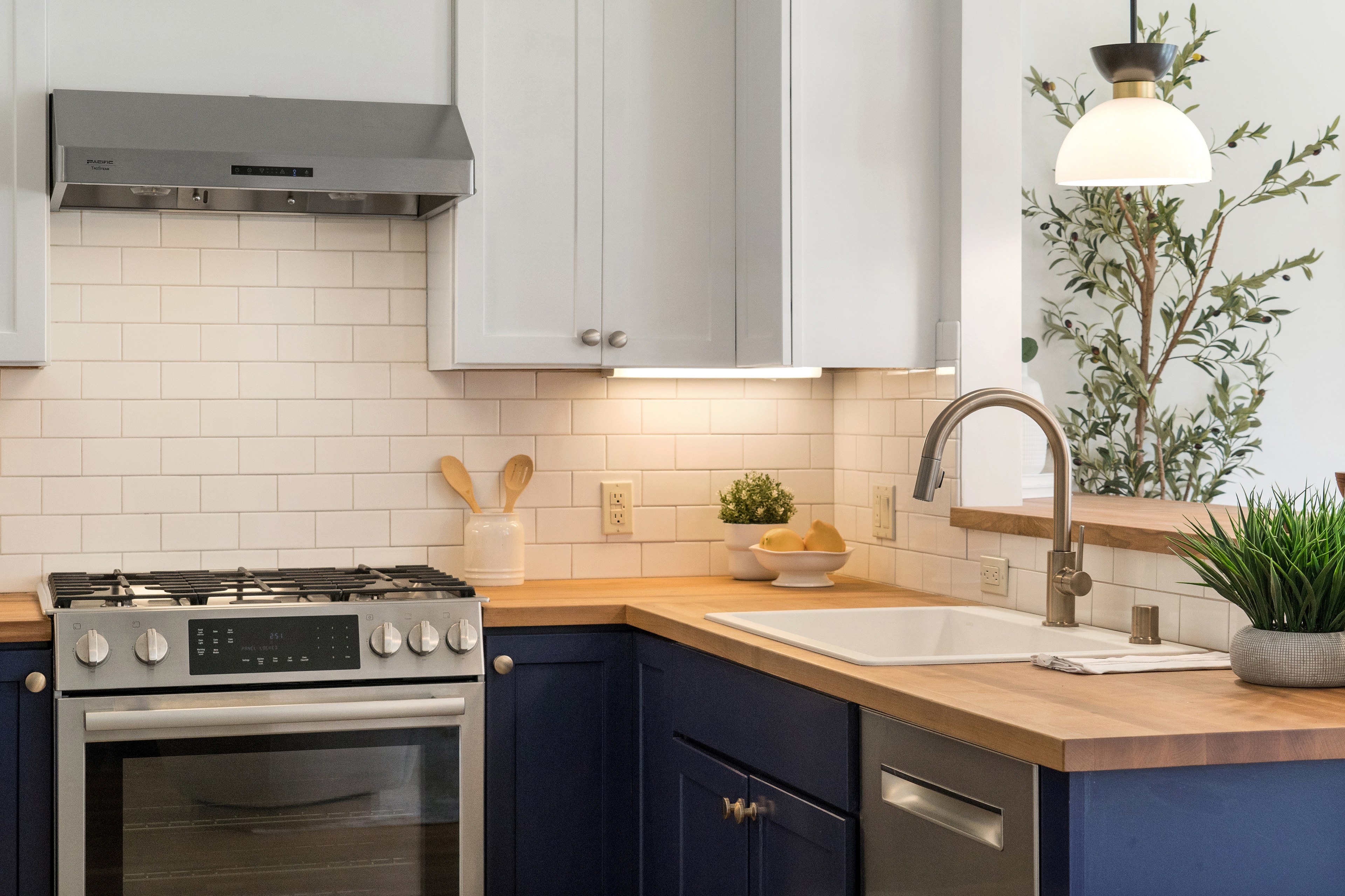 The modern kitchen features a stainless steel stove and hood, light blue cabinets, wooden countertops, white tile backsplash, potted plants, and a pendant light.