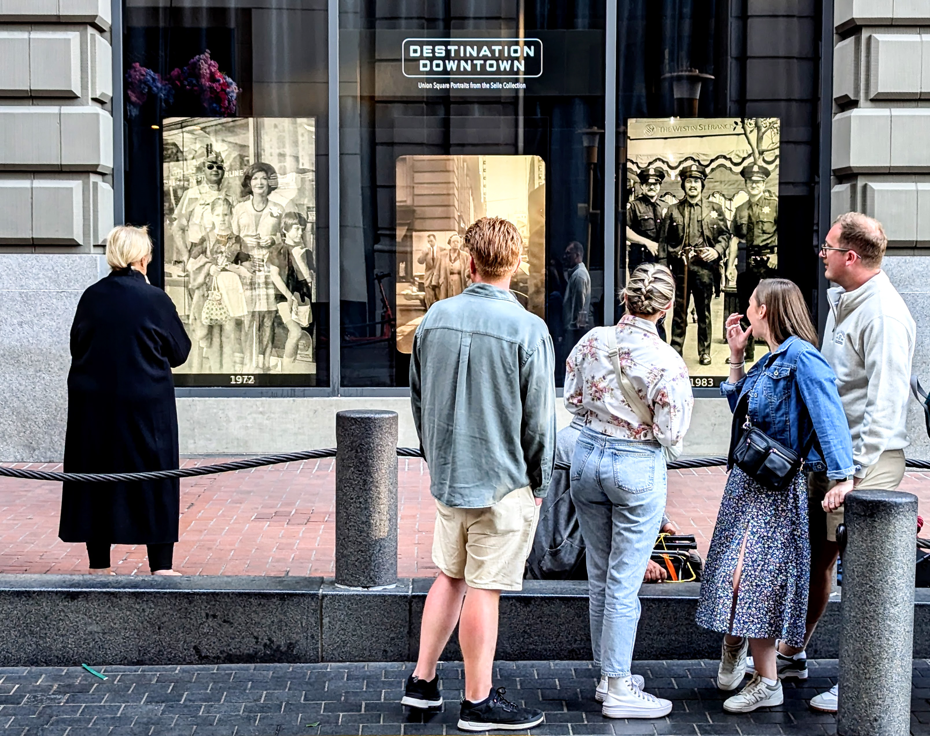 A group of people in casual clothing stand outside a building, looking at historical black-and-white photos displayed in large windows labeled &quot;Destination Downtown.&quot;