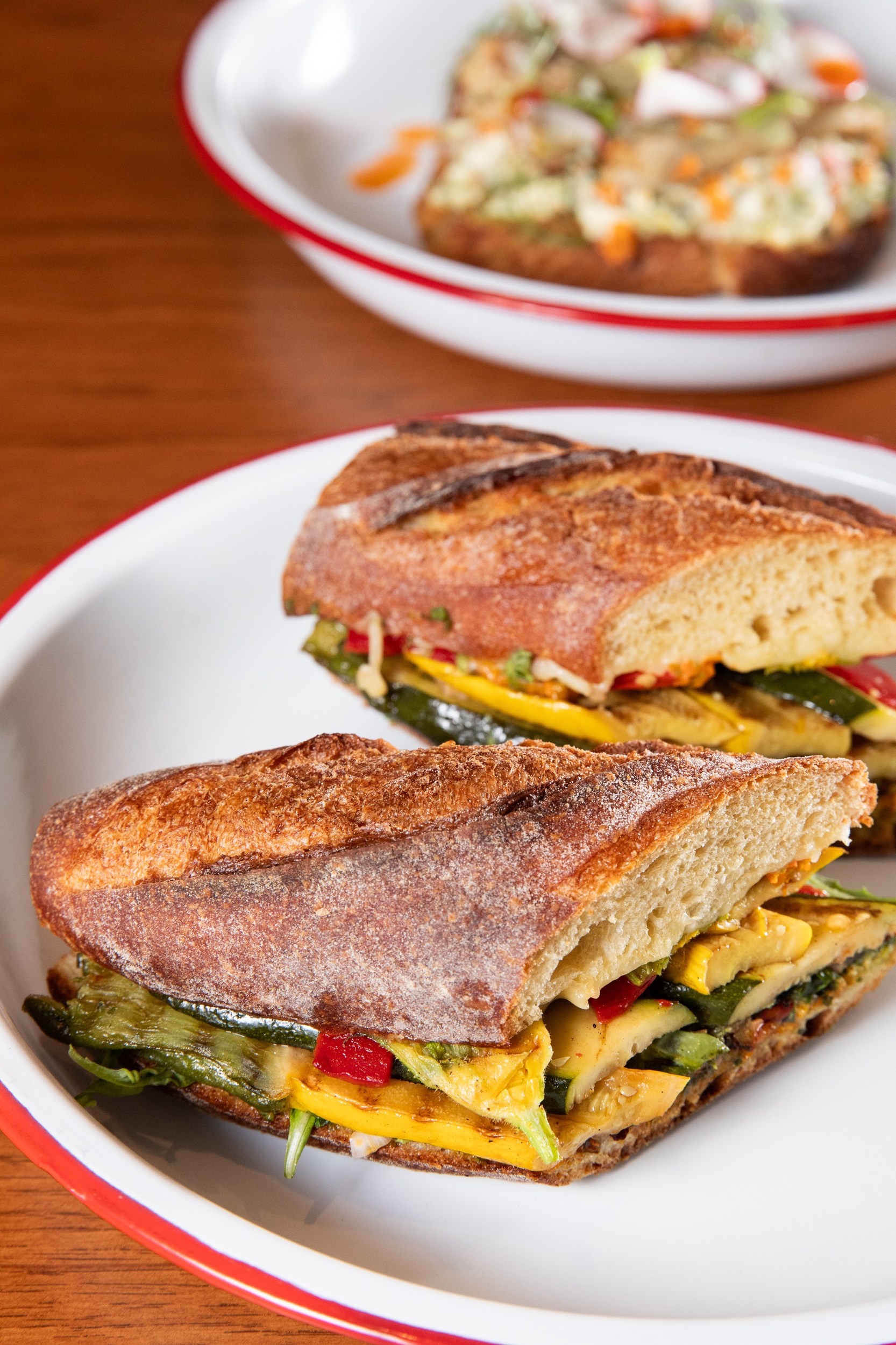 The image shows a crusty sandwich with roasted vegetables on a white plate with a red rim. In the background, there's an open-faced sandwich on a similar plate.