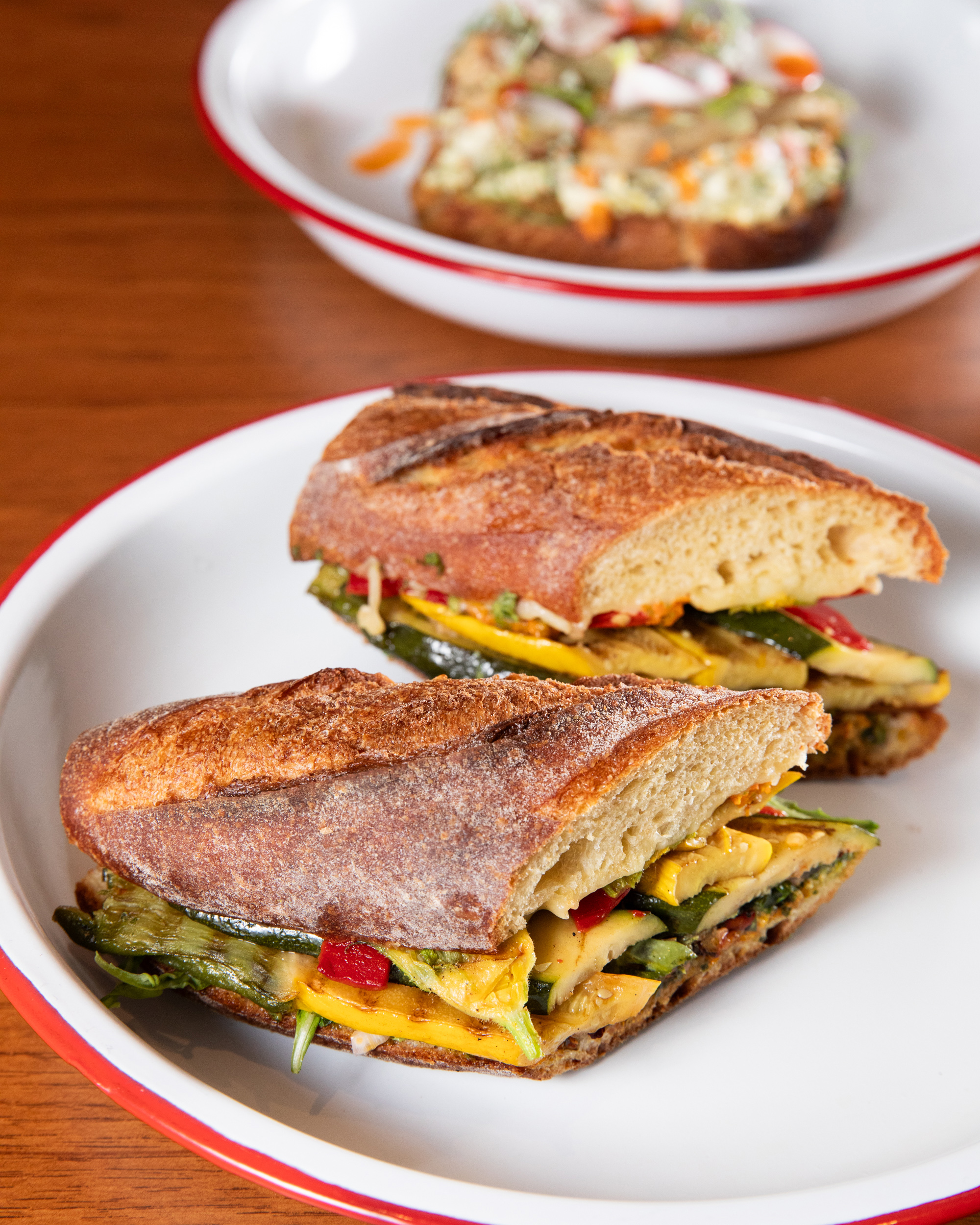 The image shows a crusty sandwich with roasted vegetables on a white plate with a red rim. In the background, there's an open-faced sandwich on a similar plate.