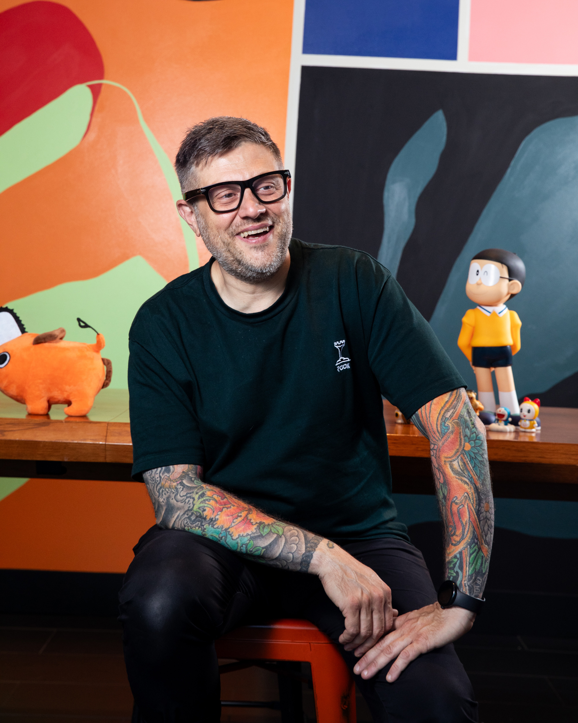 A smiling man with glasses and colorful arm tattoos sits casually on a stool. Behind him are orange plush toys and a cartoon figure, against a vibrant, abstract mural.