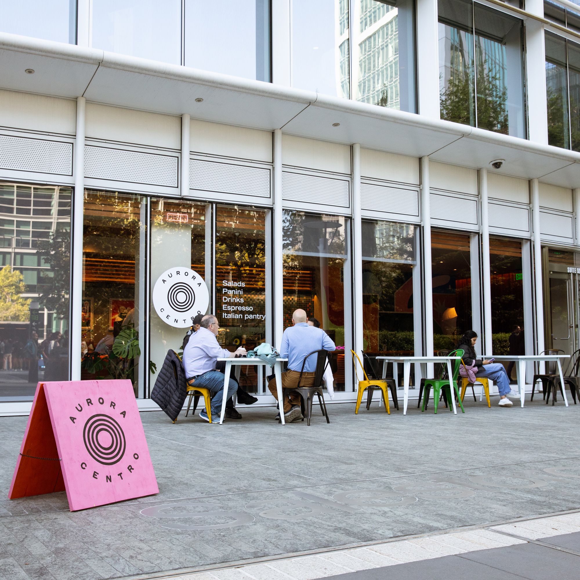 The image shows a street-level view of &quot;Aurora Centro,&quot; a cafe with a modern glass facade. Two people sit at an outdoor table, with a pink sidewalk sign nearby.