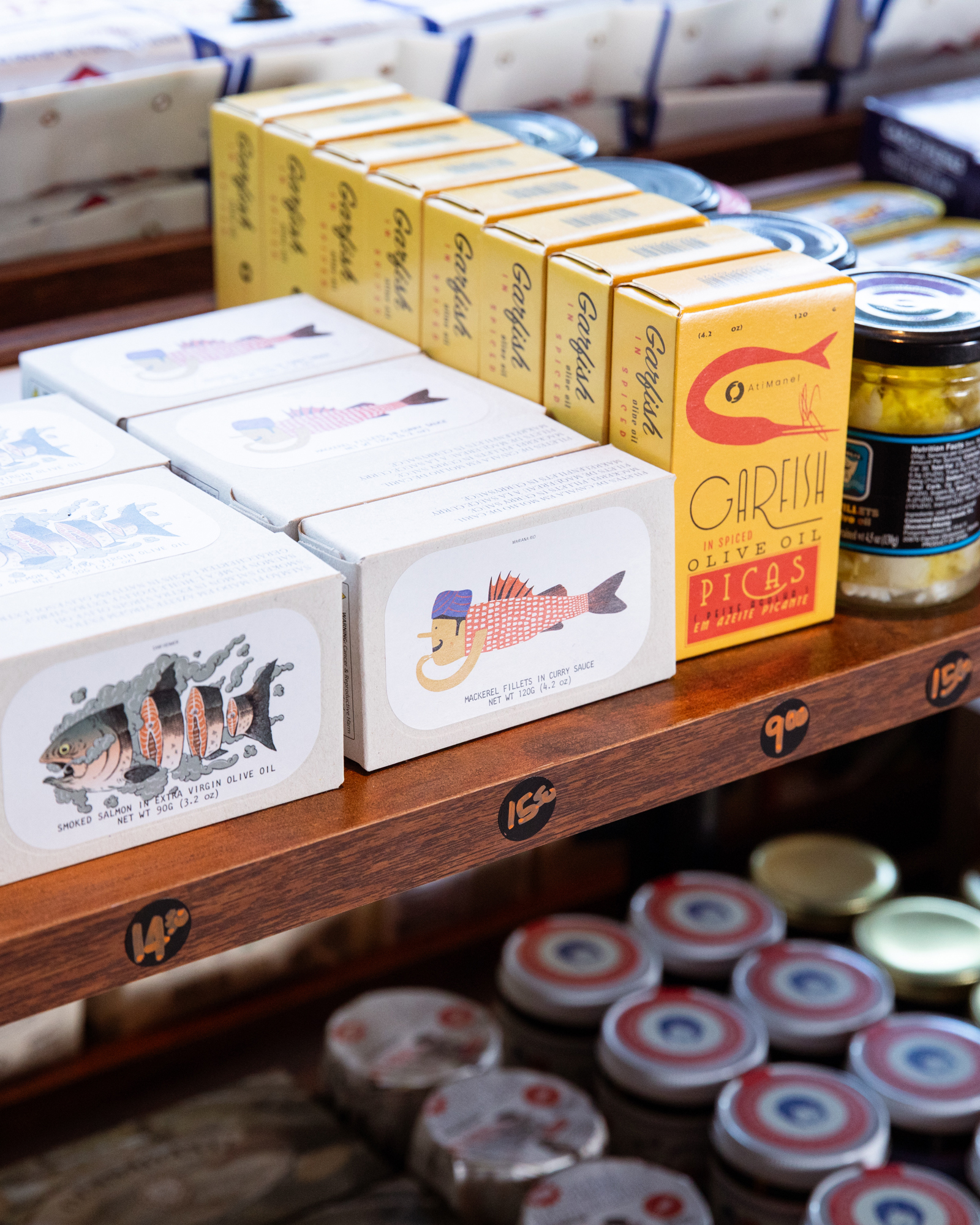 The image shows boxed sardines and mackerel fillets arranged neatly on wooden shelves, alongside jars of preserved goods, with clear price labels beneath.