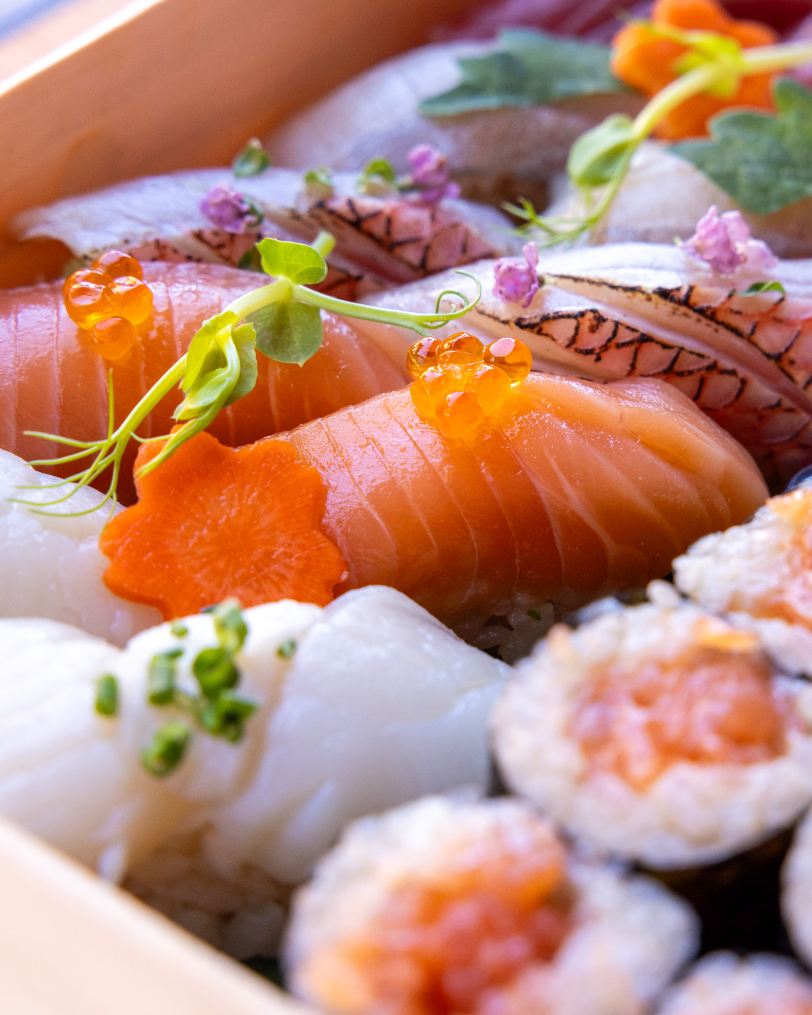 This image shows a variety of sushi pieces, including slices of salmon, topped with fish roe, a small orange flower cutout, and some green garnishes.